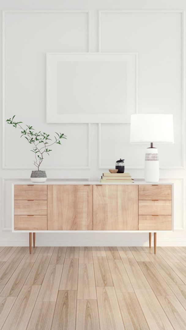 Un buffet en bois décoré sous un cadre photo blanc vierge sur un mur blanc ; parquet clair.