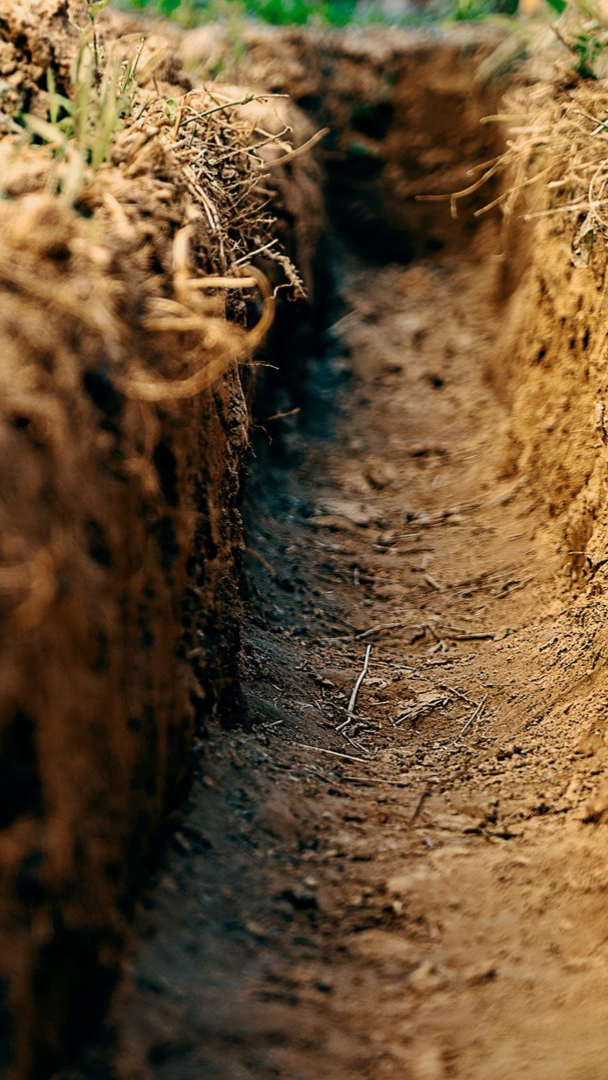 Tranchée étroite creusée dans la terre brune, bordée d'herbe sur un côté.