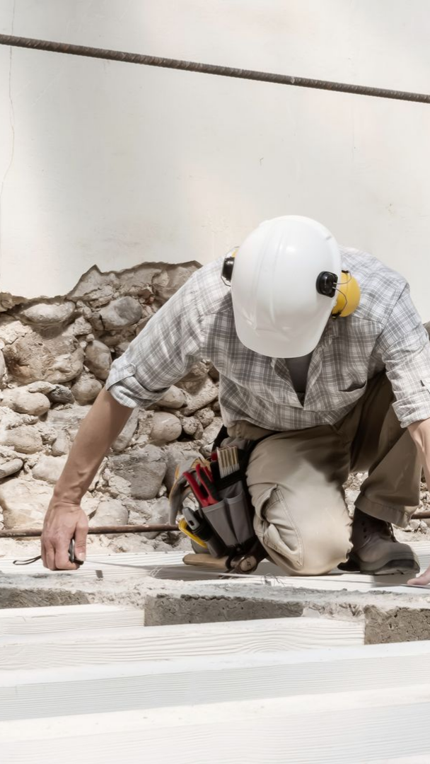 Un ouvrier du bâtiment examine une surface en béton, portant un casque de chantier et une ceinture porte-outils.