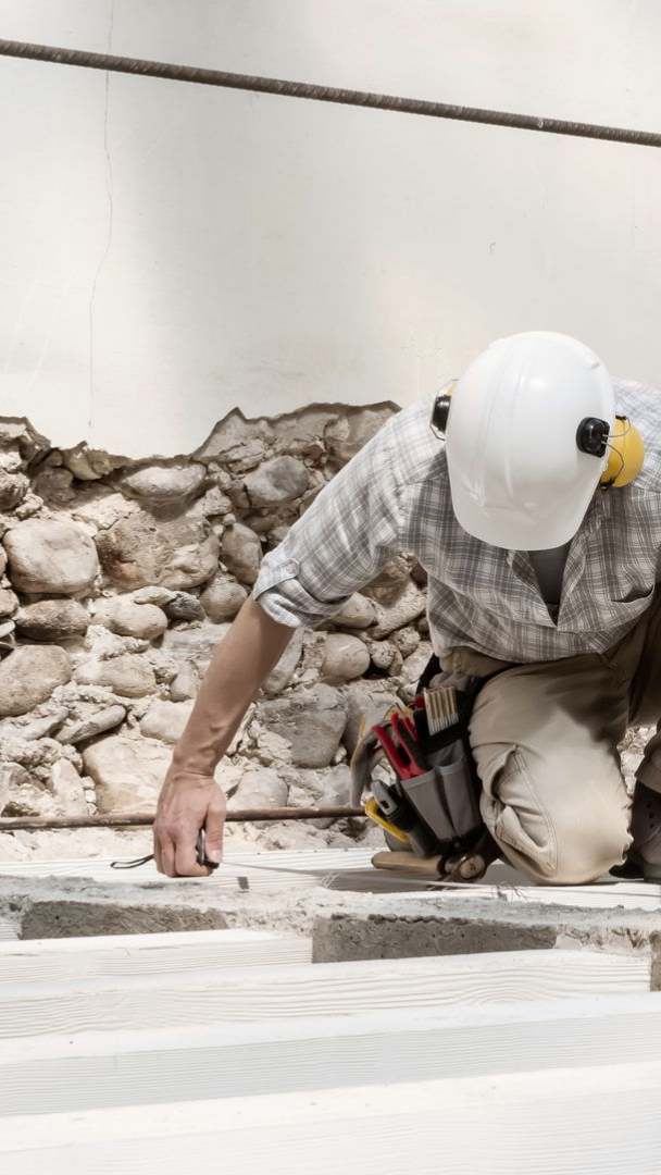 Ouvrier du bâtiment à genoux, inspectant des blocs de béton ; casque blanc, ceinture porte-outils.