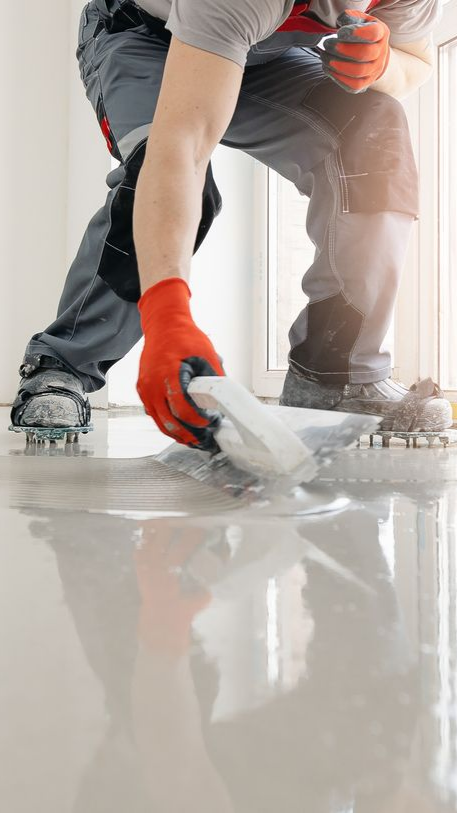 Une personne nivelant un sol gris et humide à l'aide d'une truelle, portant des gants rouges et des vêtements de travail.