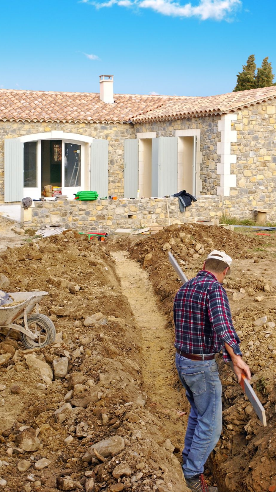 Une tranchée devant une maison en pierre aux volets bleu clair, sous un ciel bleu.
