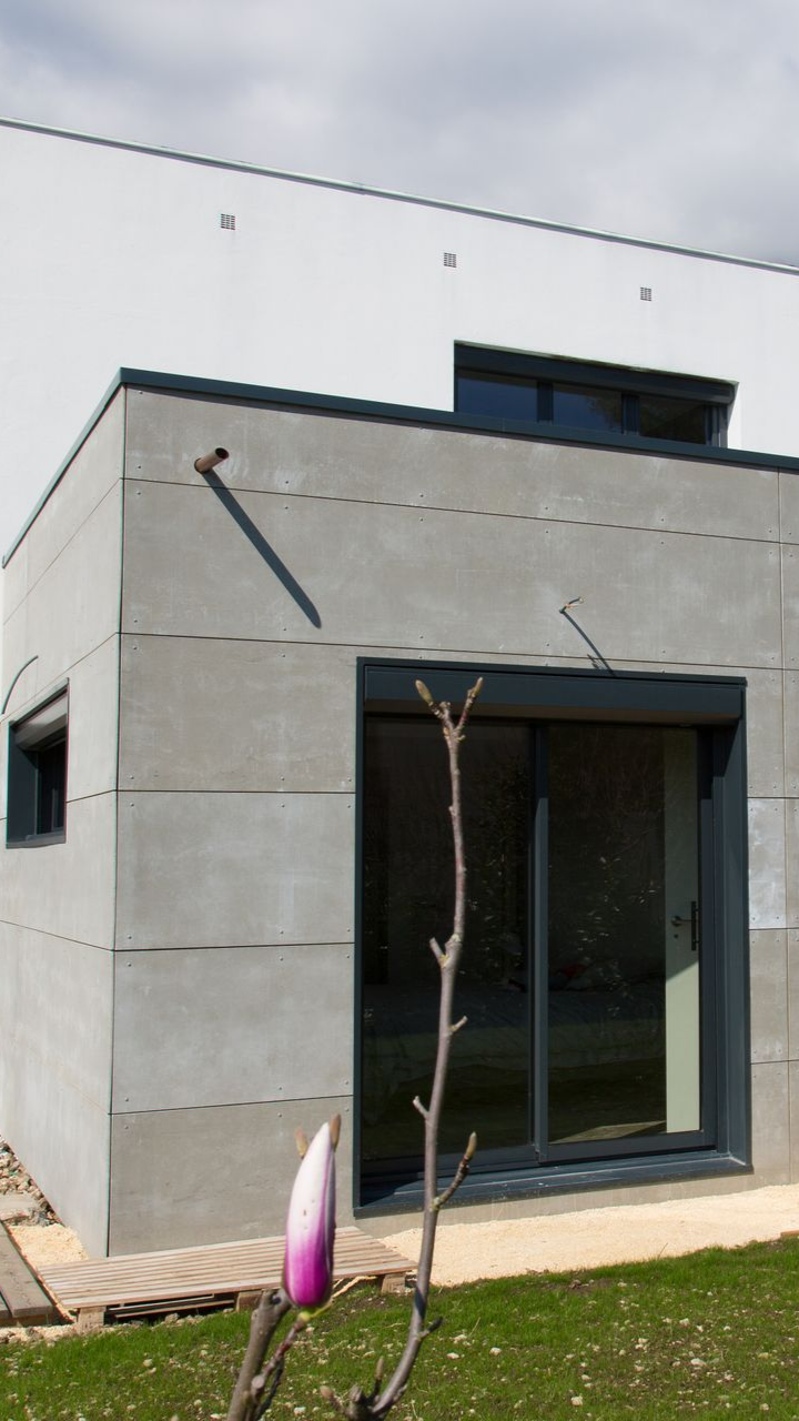 Bâtiment revêtu de béton gris, avec des encadrements de fenêtres sombres, une petite fenêtre et une porte coulissante en verre.