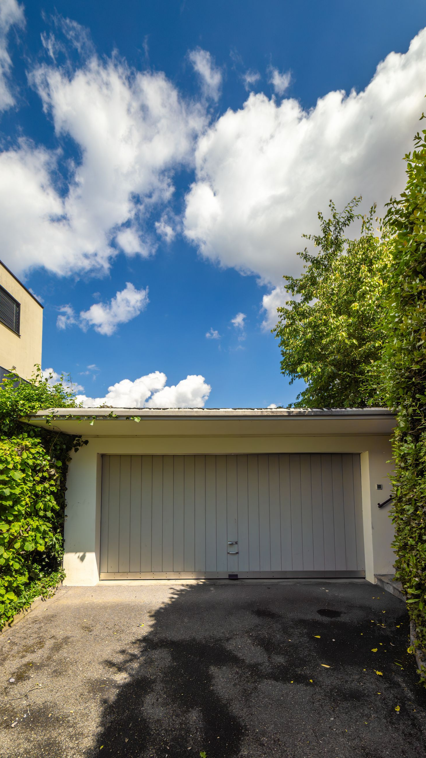 Garage blanc sous un ciel bleu parsemé de nuages cotonneux. Allée sombre, végétation verte de part et d'autre.