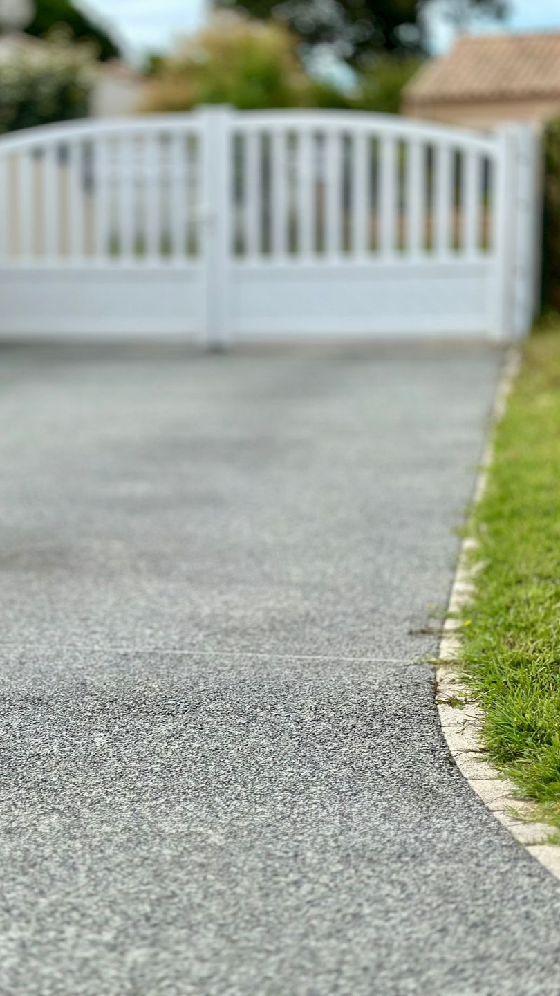 Allée de gravier gris menant à un portail blanc ; l'allée est bordée d'herbe verte.