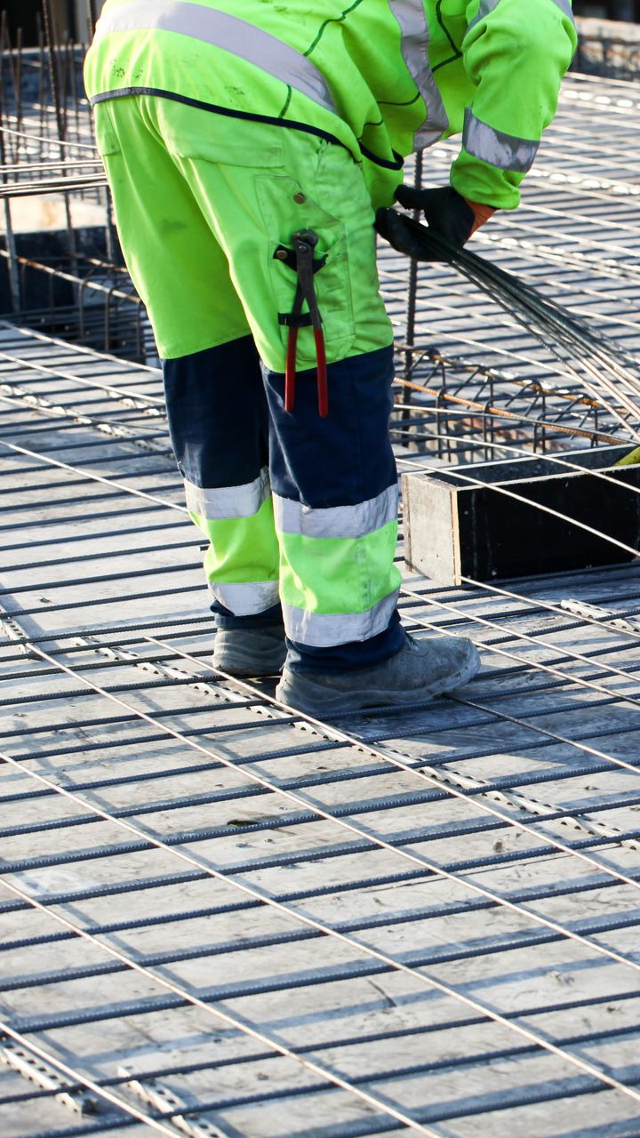 Un ouvrier du bâtiment portant un équipement de sécurité travaille avec des barres d'armature sur une dalle de béton, à l'aide d'une pince.