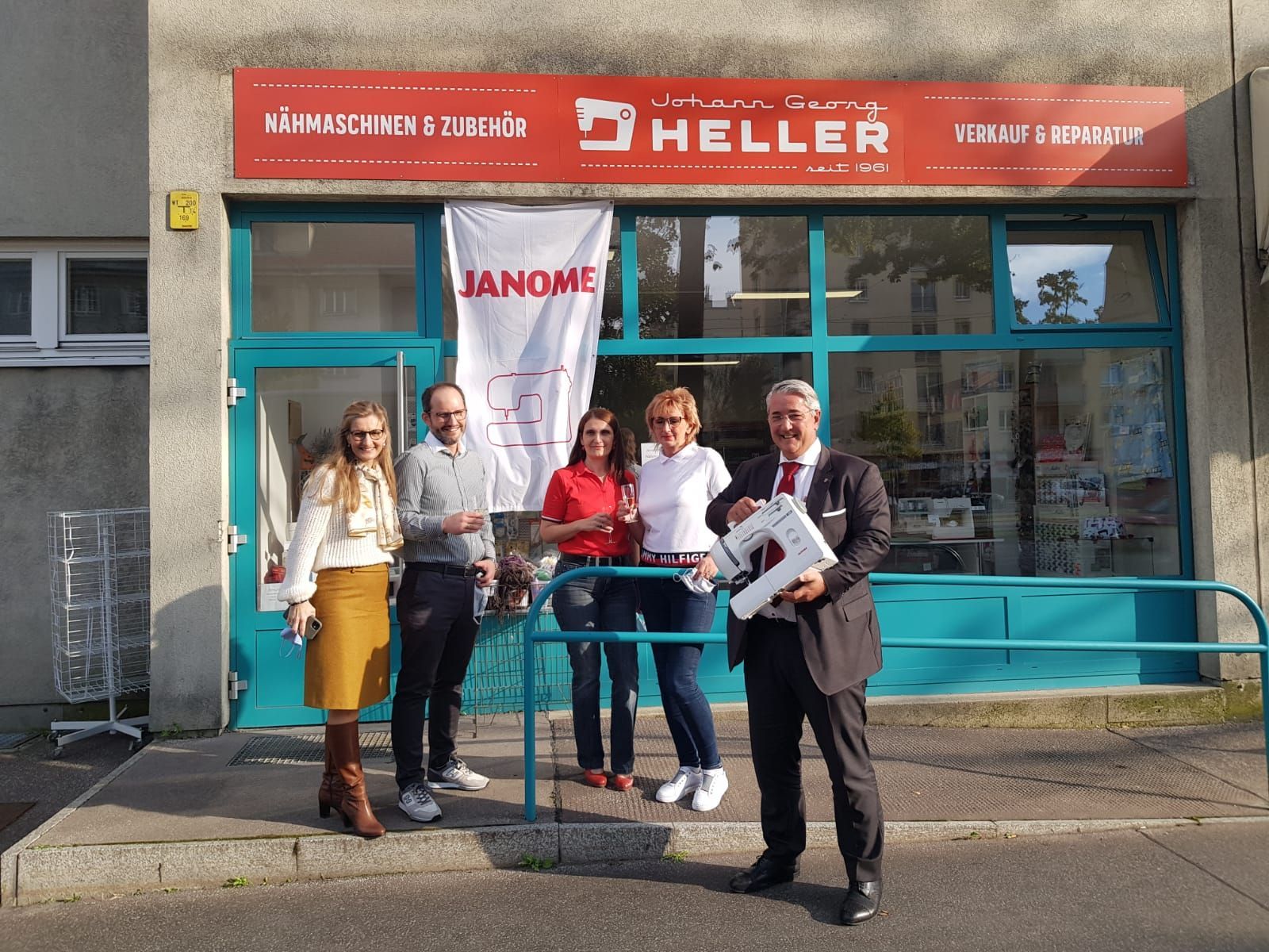 Eine Gruppe von Personen posiert vor einem Geschäft mit Nähmaschine. Das Geschäft hat ein Heller-Schild und Janome-Banner.
