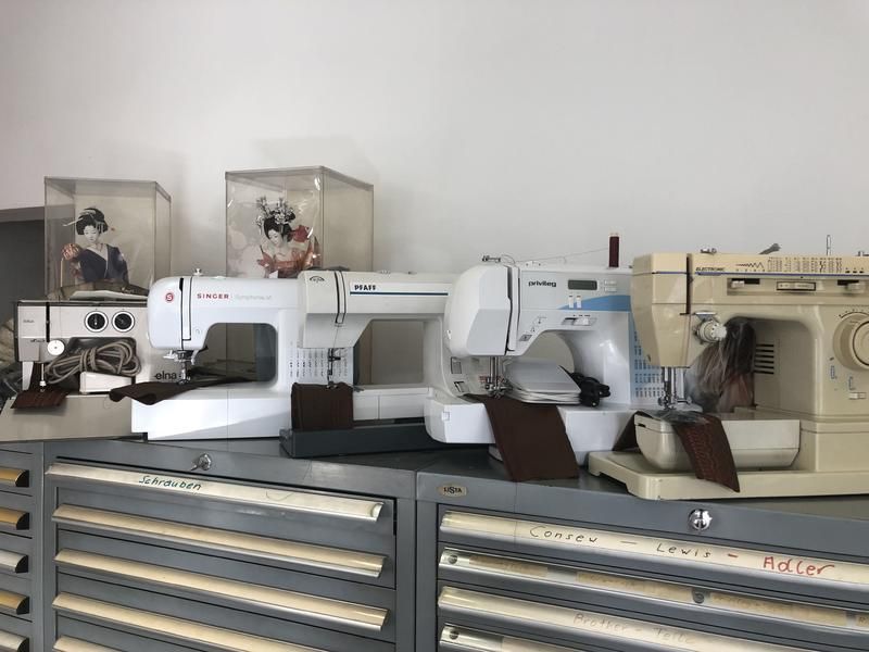 Nähmaschinen auf einem Regal mit Schubladen, eine weiße Wand und Schaukartons mit Puppen.