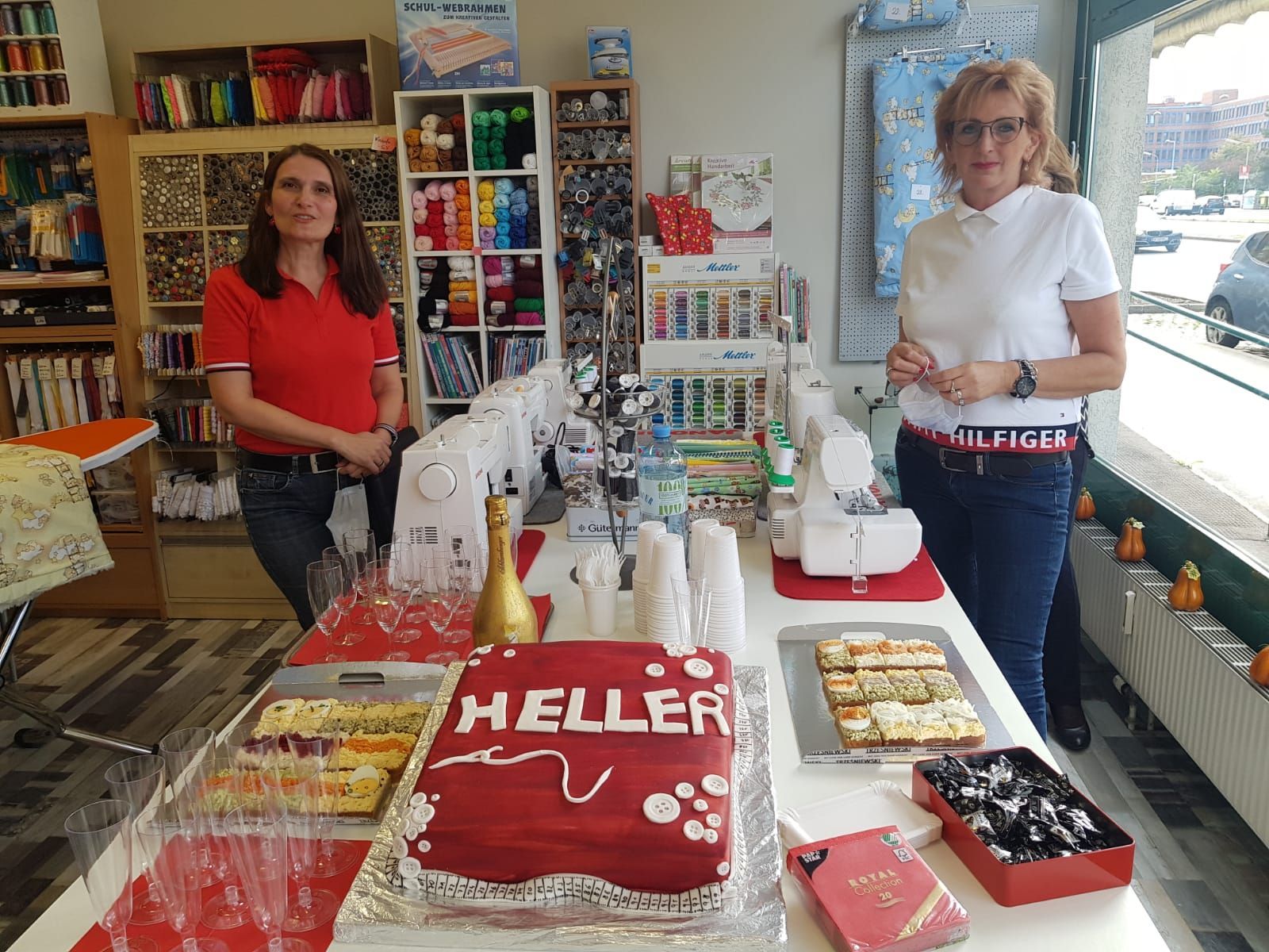 Zwei Frauen stehen hinter einem Tisch mit einer Torte mit der Aufschrift „Heller“, Snacks und Nähmaschinen.