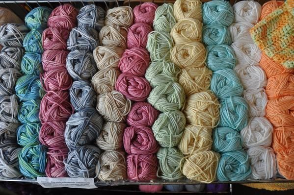 Reihen bunter Garnstränge auf Regalen; darunter Rosa-, Blau-, Gelb- und Cremetöne.