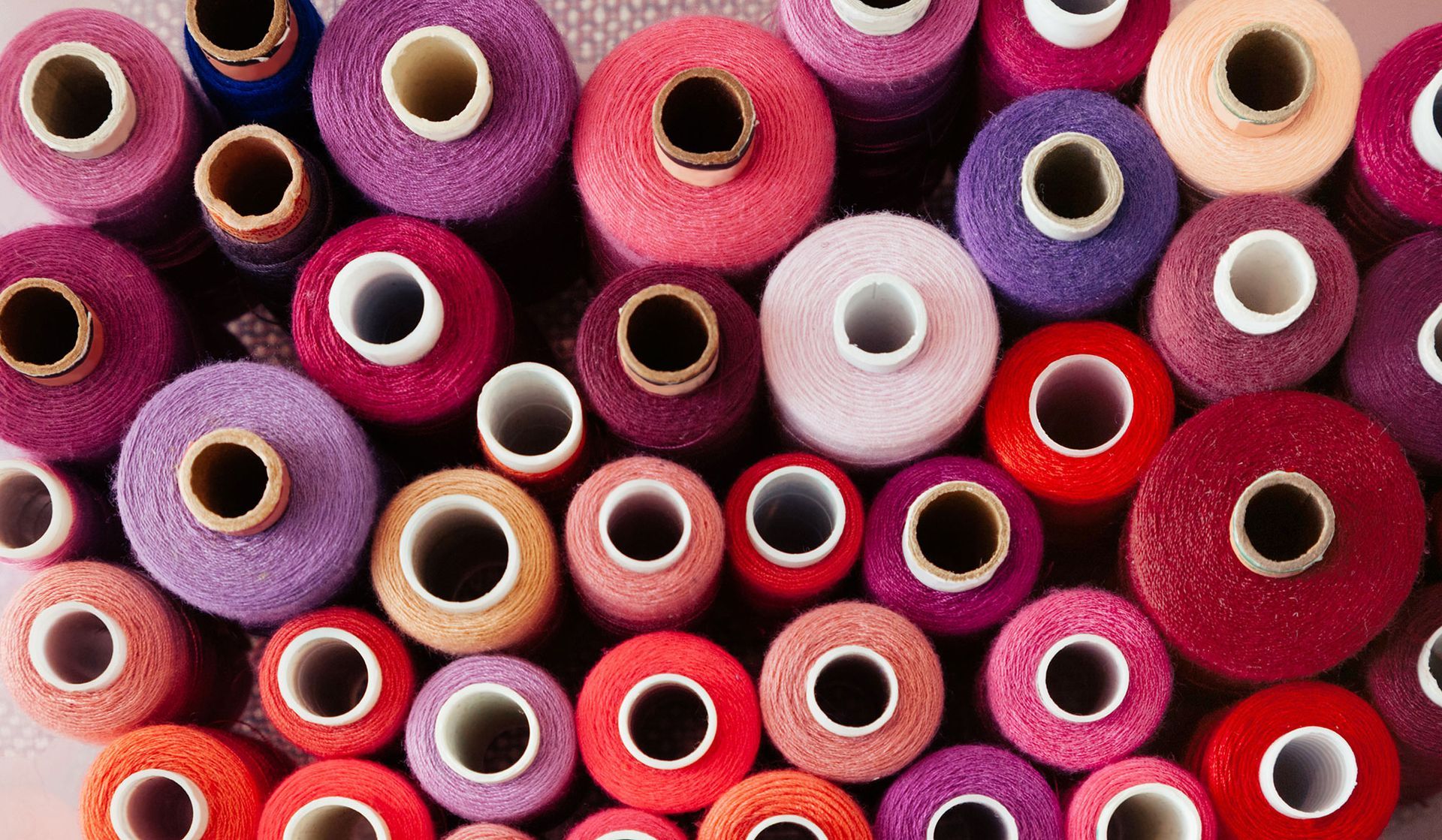 Spulen mit buntem Garn, hauptsächlich in Rosa-, Lila- und Rottönen, dicht beieinander angeordnet.