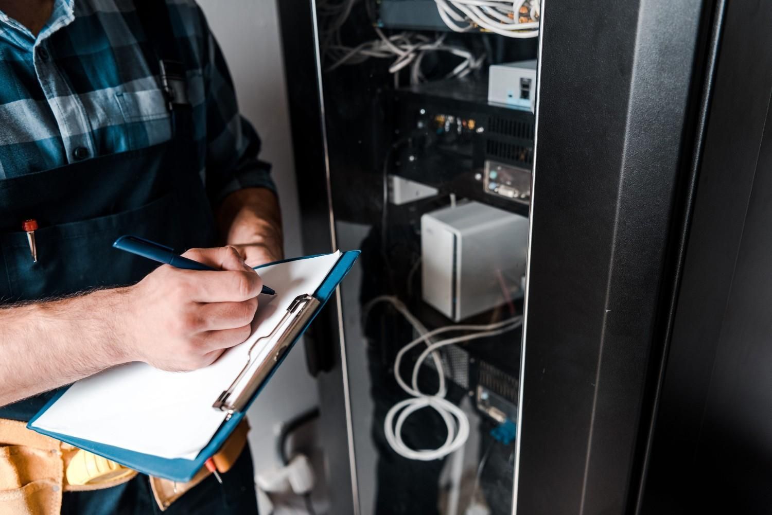 Técnico escribiendo en un portapapeles junto a un rack de servidores abierto con cables y equipos.
