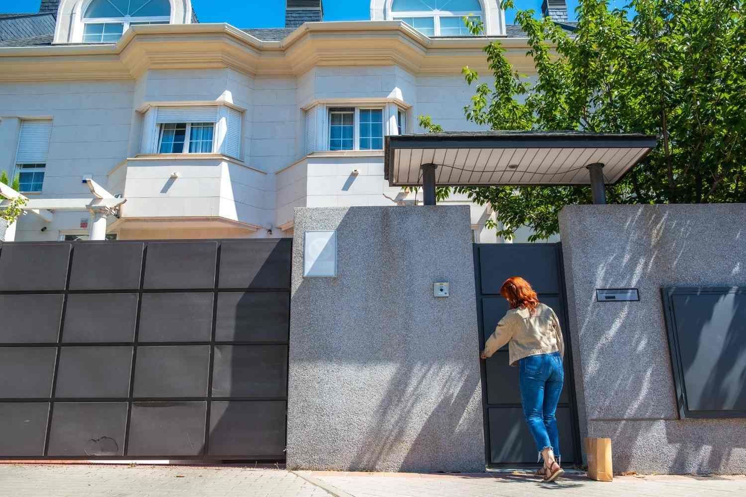 Una persona abre una puerta gris frente a un edificio azul claro y un muro de piedra.