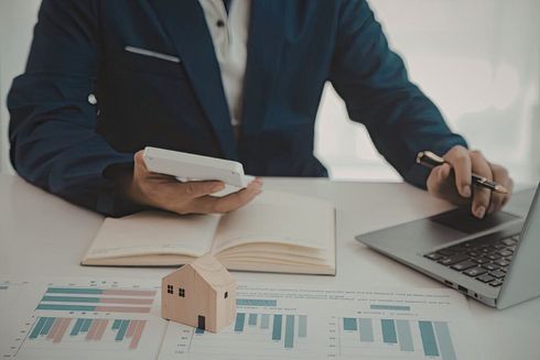 Persona revisando documentos financieros en un escritorio con gráficos, una computadora portátil, una calculadora y una pequeña maqueta de una casa.