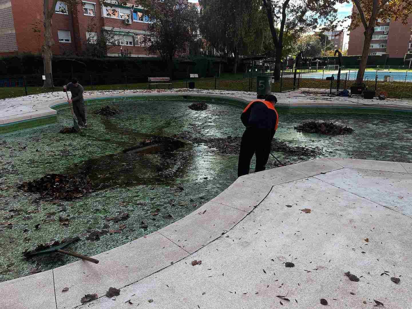 Persona vestida con ropa oscura y chaleco naranja de pie junto a un sendero embarrado e inundado en un parque.