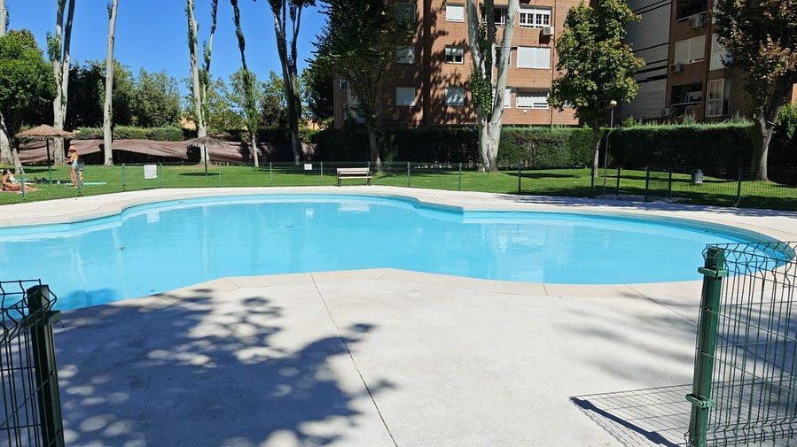 Piscina al aire libre con agua azul, terraza blanca y edificios de apartamentos y palmeras al fondo.