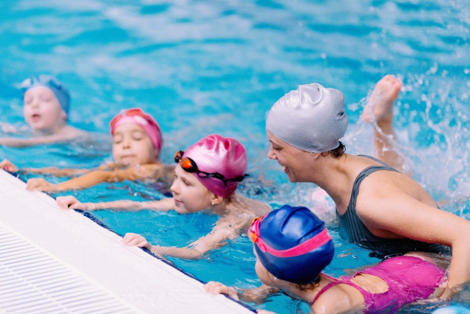 Niños con gorros de natación de colores practicando en el borde de la piscina con un entrenador cerca.