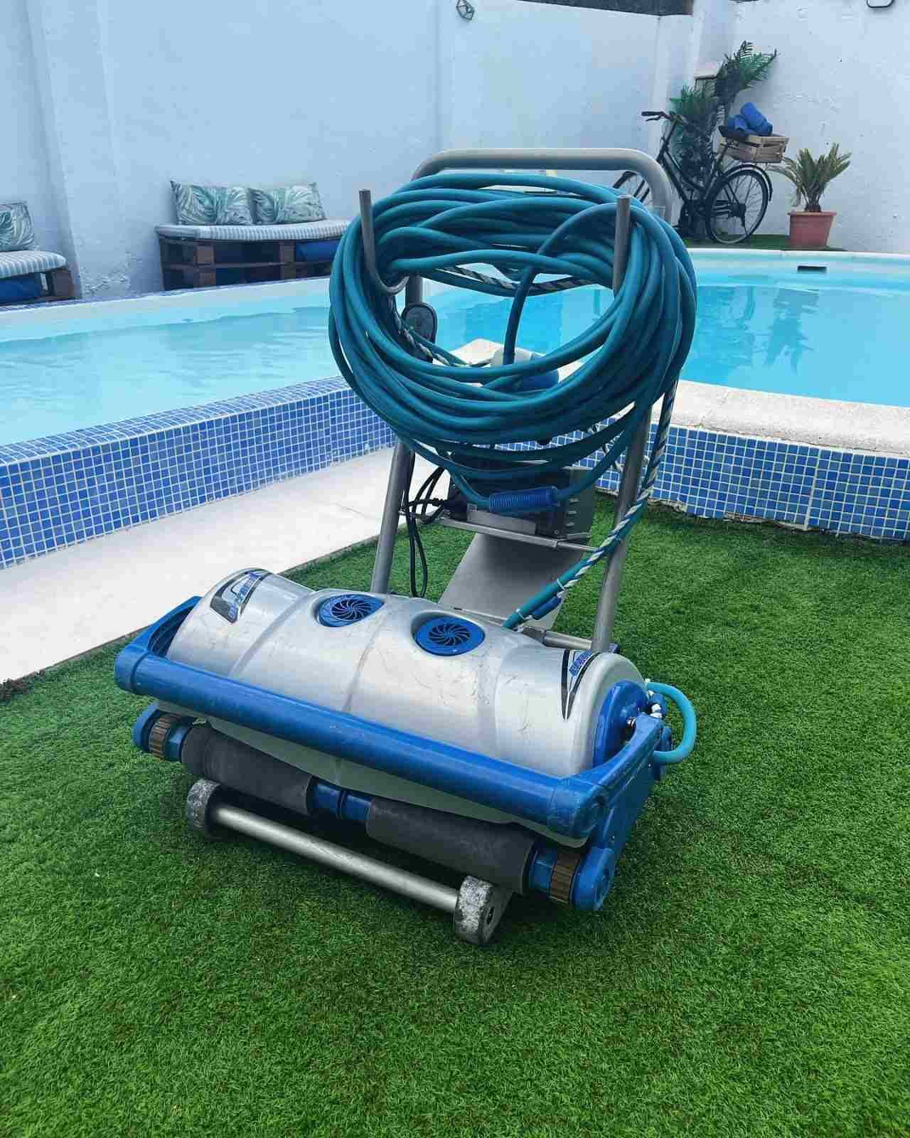 Robot limpiador de piscinas con manguera azul enrollada sobre el césped junto a una piscina.