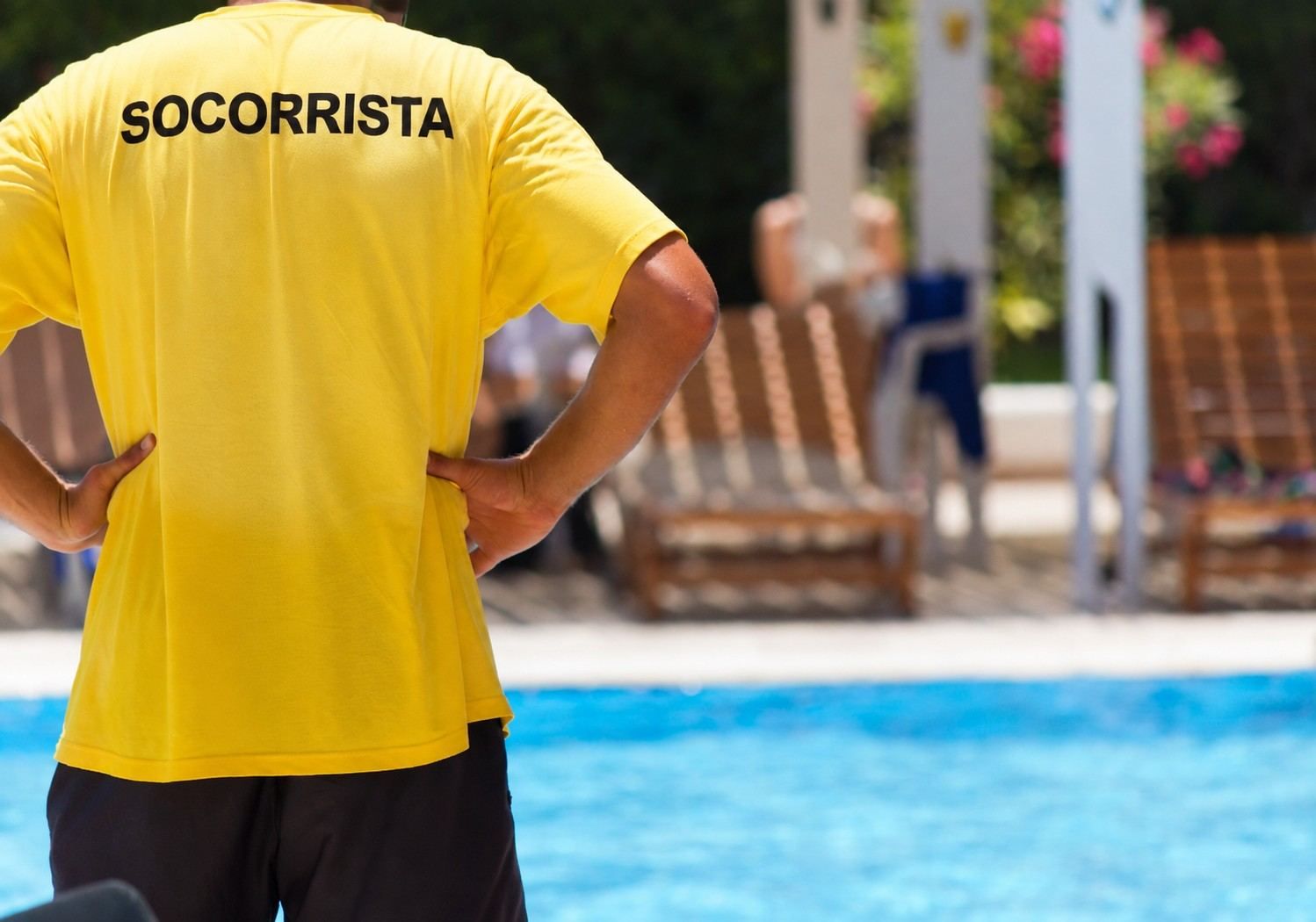 Un socorrista con una camisa amarilla está de pie junto a una piscina con tumbonas al fondo.