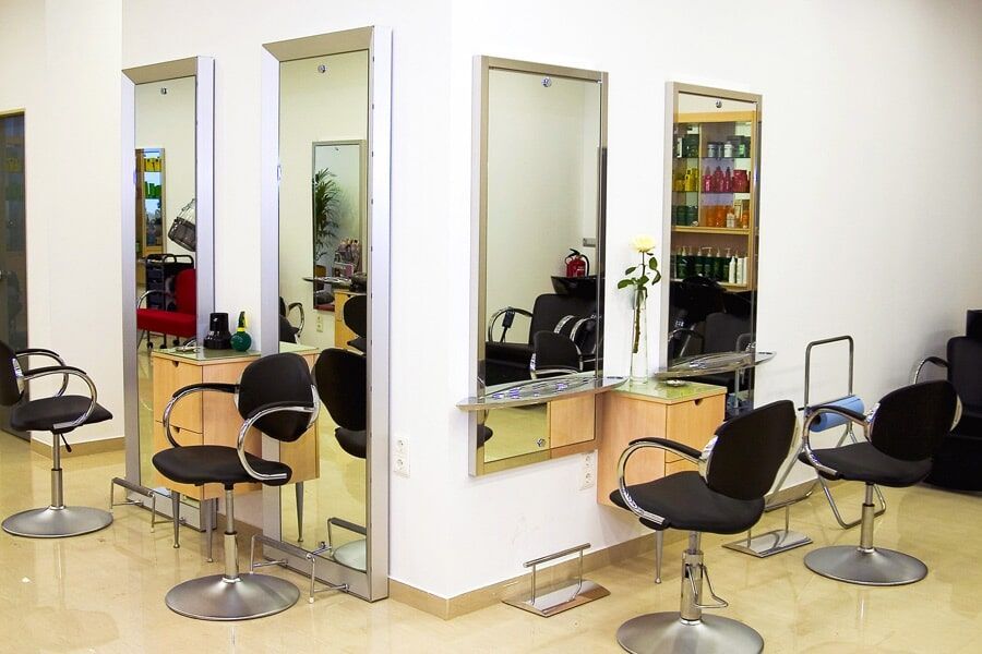 Interior de salão de cabeleireiro com espelhos, estações de trabalho e cadeiras. Ambiente luminoso, tons neutros.