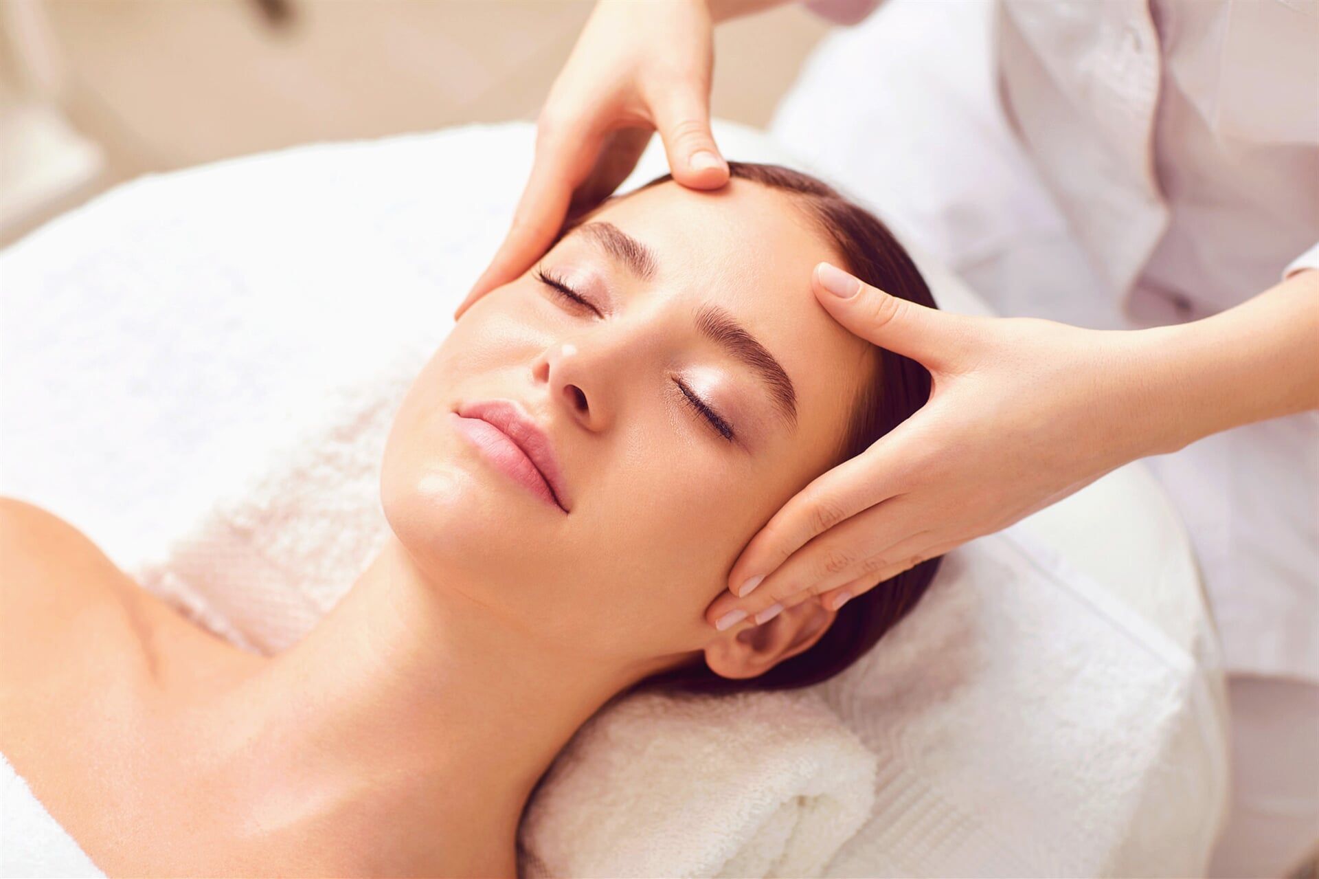 Pessoa recebendo uma massagem facial com as mãos na testa, em uma mesa e lençóis brancos.