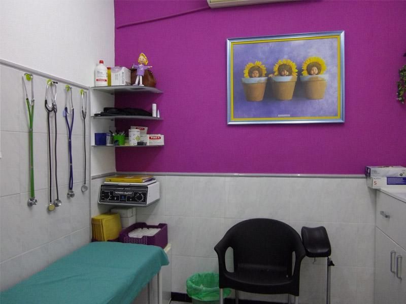 Pequeña sala de exploración con una camilla color turquesa, silla negra, estantes en la pared y una pared decorativa morada con un cuadro de flores.
