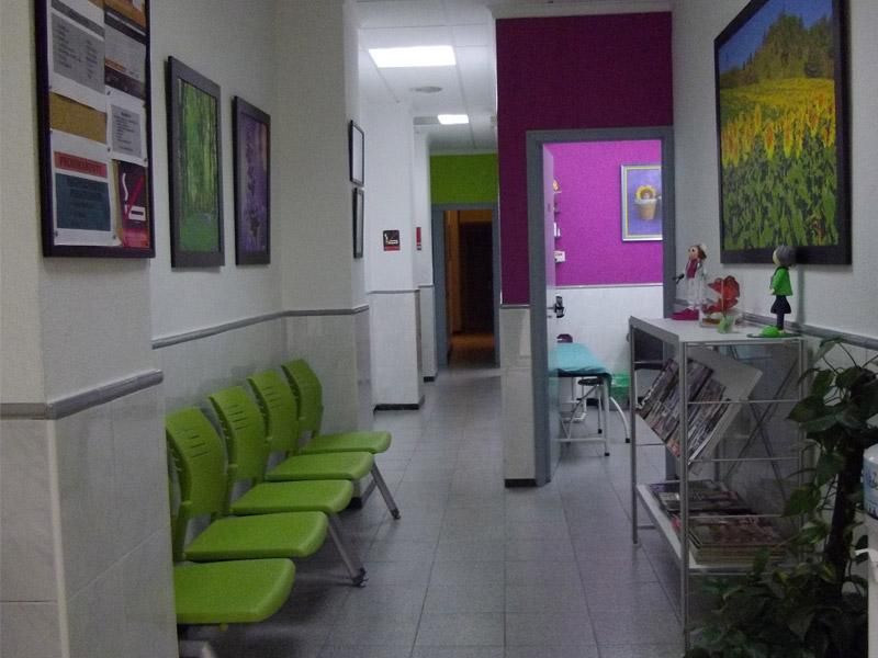 Pasillo de la clínica con sillas verdes, cuadros enmarcados y un mostrador de recepción bajo una pared de color púrpura.