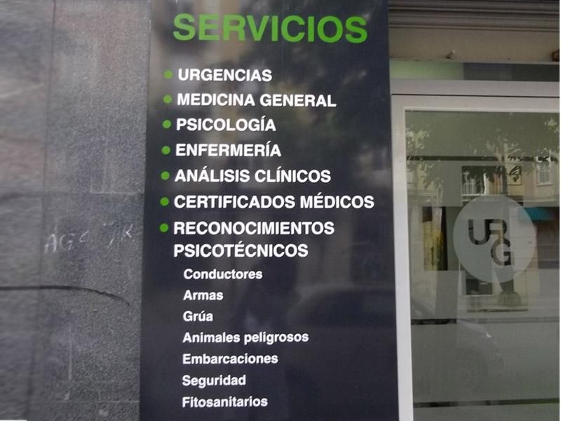 Servicios médicos y psicotécnicos en un cartel negro con texto verde y blanco, junto a una puerta de vidrio.