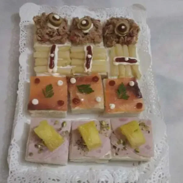 Una bandeja rectangular con una variedad de sándwiches pequeños rellenos de atún, aceitunas, salmón, piña y espárragos.