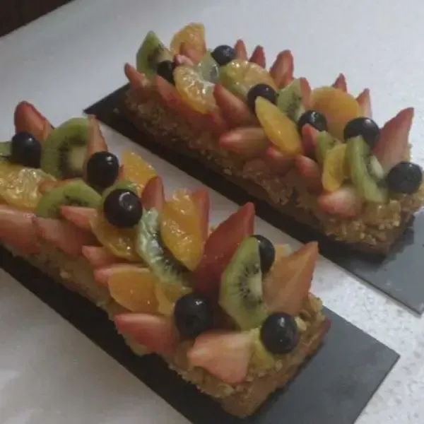 Dos tartaletas rectangulares de frutas cubiertas con fresas en rodajas, kiwi, gajos de mandarina y arándanos