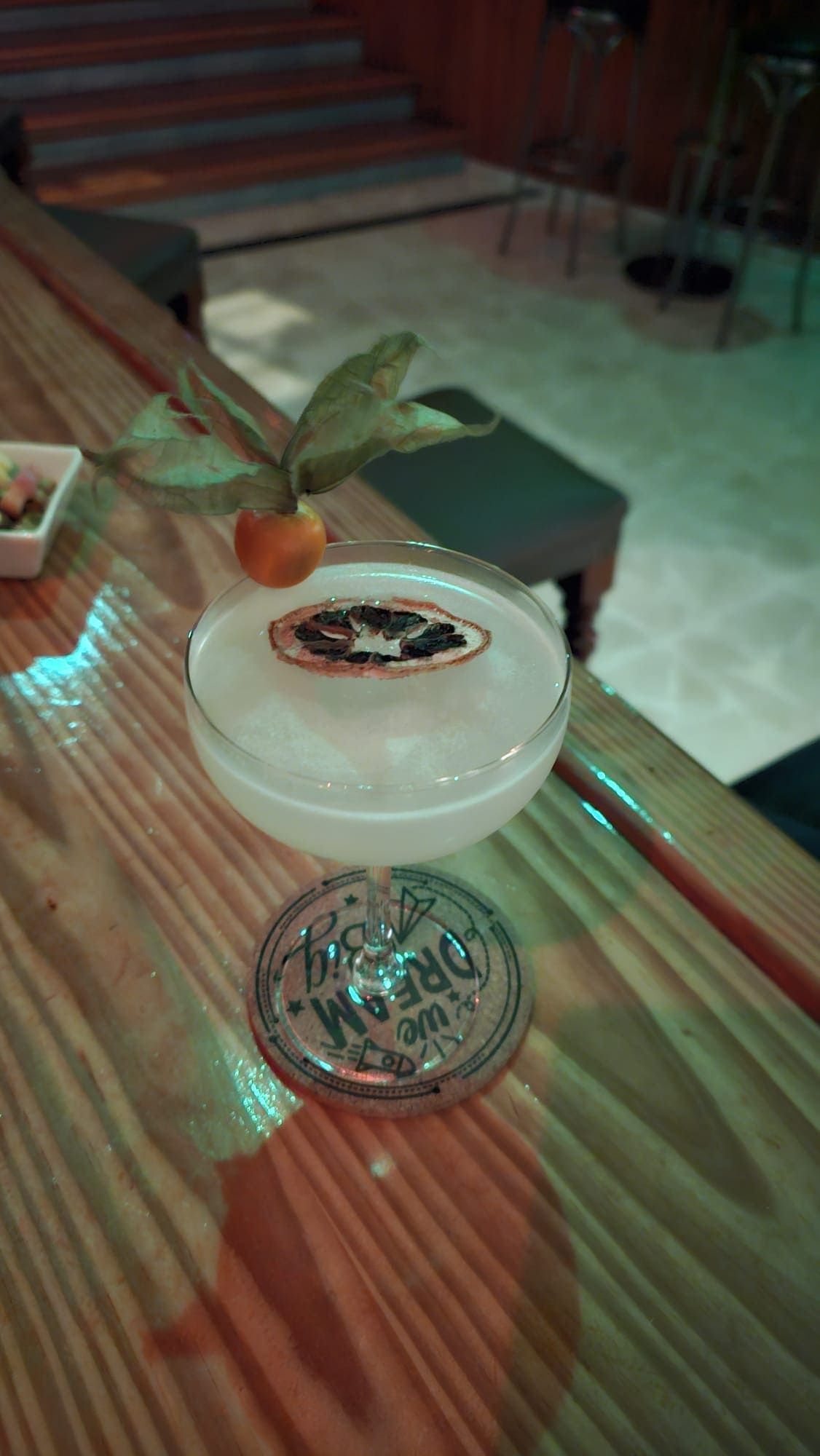 Un cóctel servido en una copa con tallo, coronado con una rodaja de cítricos secos y una baya de physalis, sobre una barra de madera.