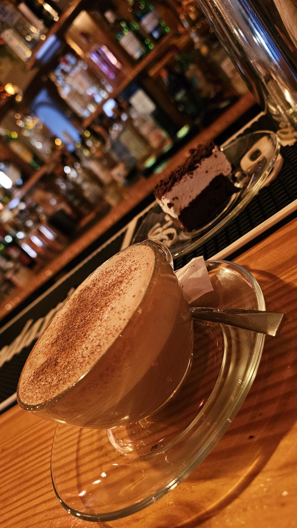 Una taza de café con espuma y cacao en polvo, junto a una pequeña porción de pastel, sobre una barra de madera.