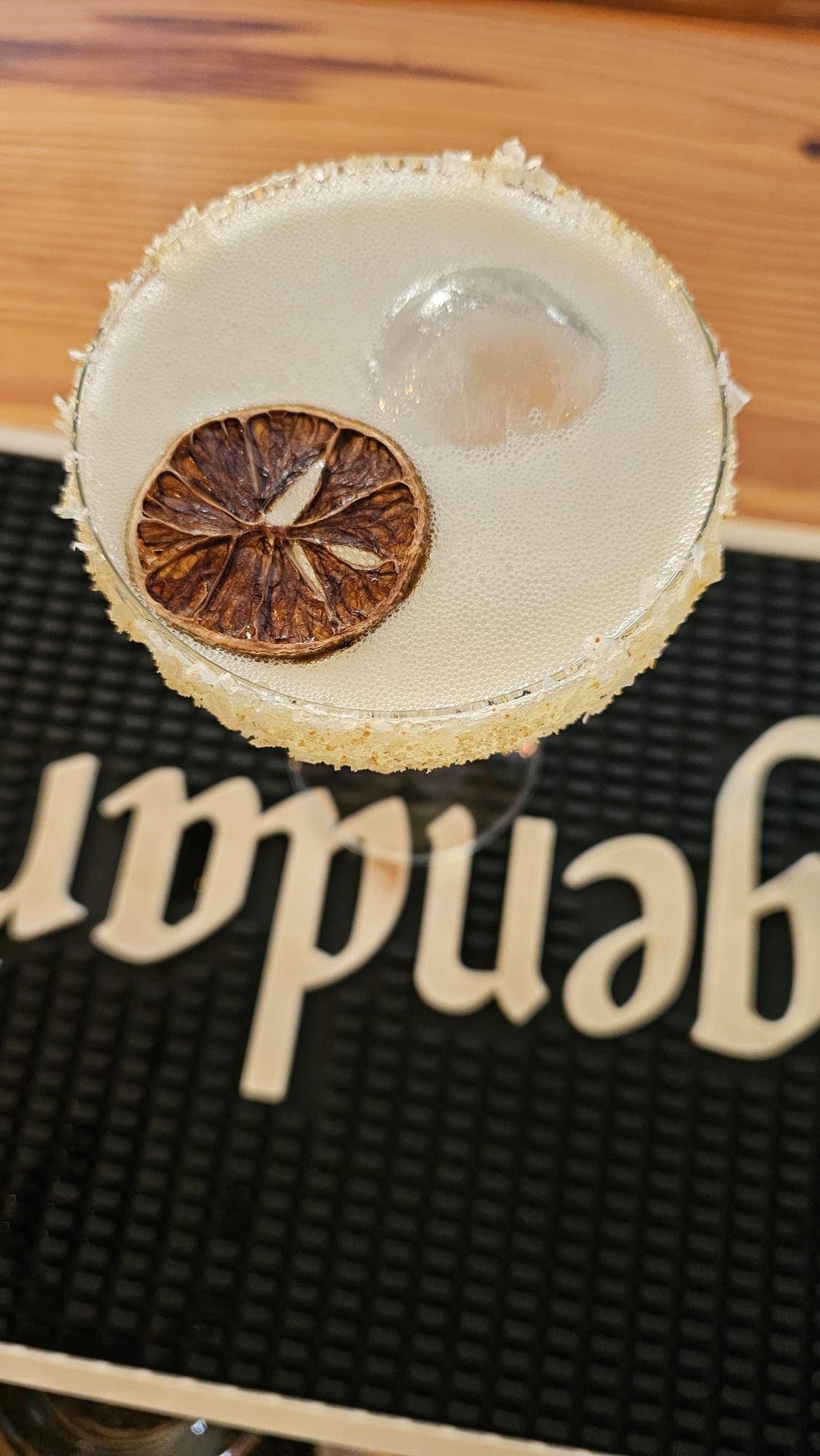 Un cóctel pálido con el borde escarchado con sal, un cubo de hielo grande y redondo, y una rodaja de lima deshidratada como guarnición sobre un tapete de bar negro.