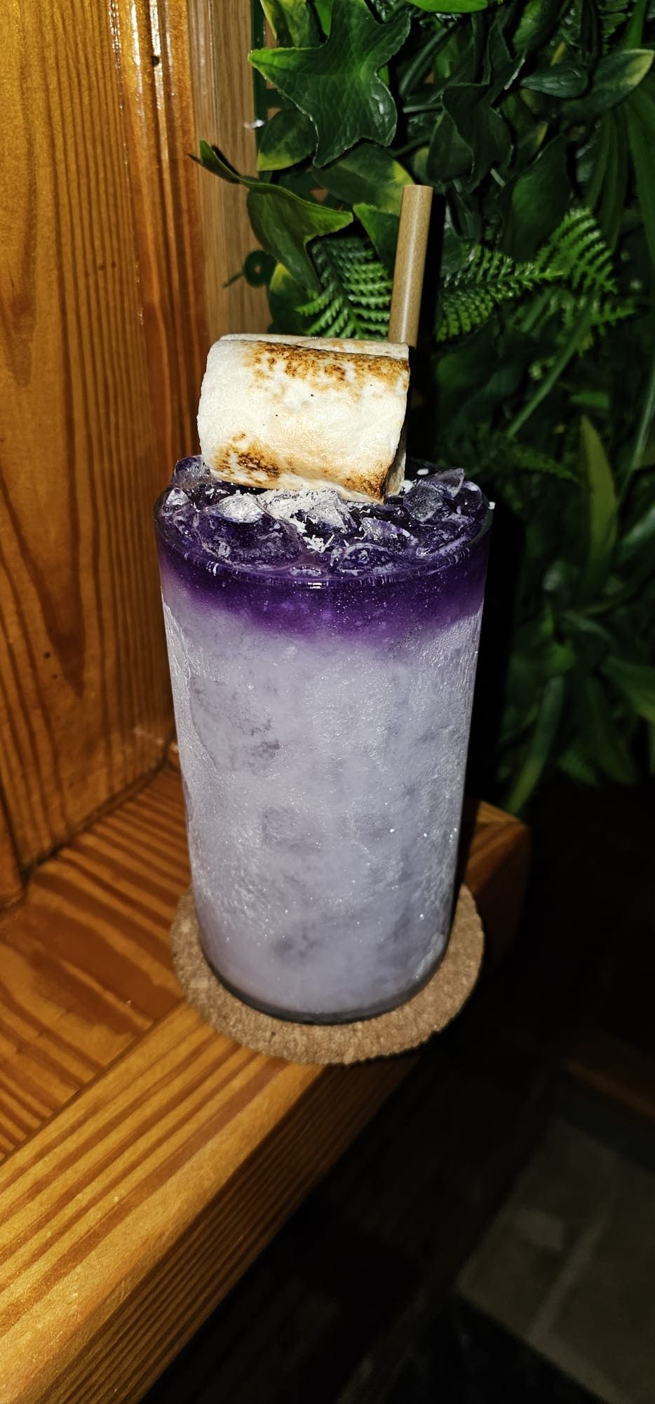 Un cóctel de color púrpura intenso servido en un vaso escarchado con hielo, coronado con un malvavisco tostado y colocado sobre un posavasos.