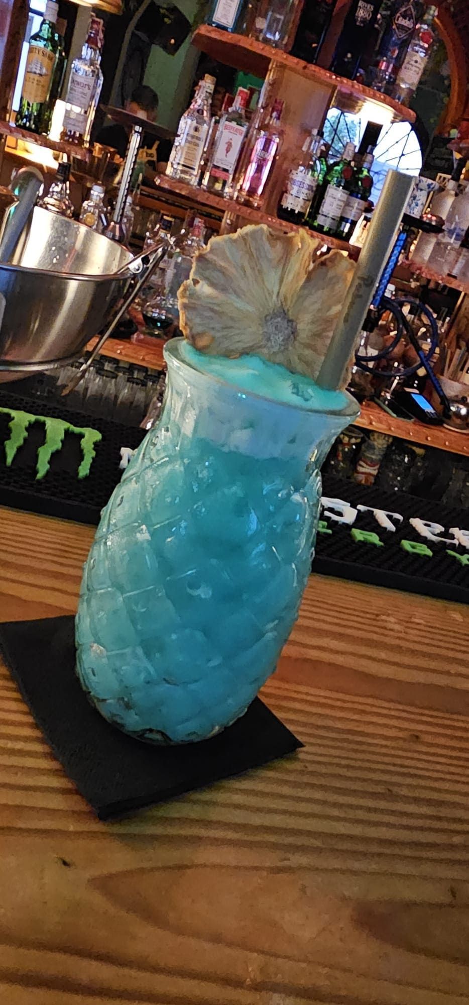Un cóctel tropical de color azul brillante servido en un vaso con forma de piña, adornado con una rodaja de piña deshidratada sobre una barra de madera.