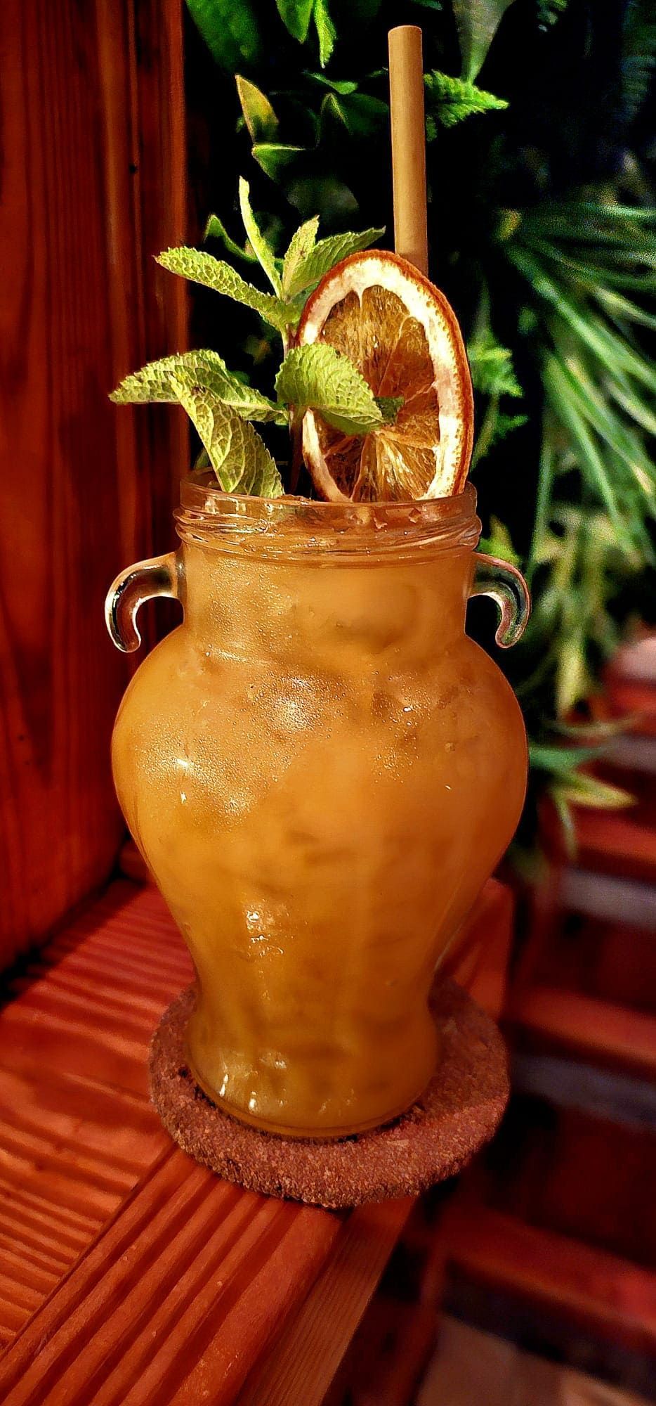 Un cóctel de color ámbar en un jarrón de cristal con dos asas, adornado con una rodaja de naranja deshidratada y menta sobre un posavasos de corcho.