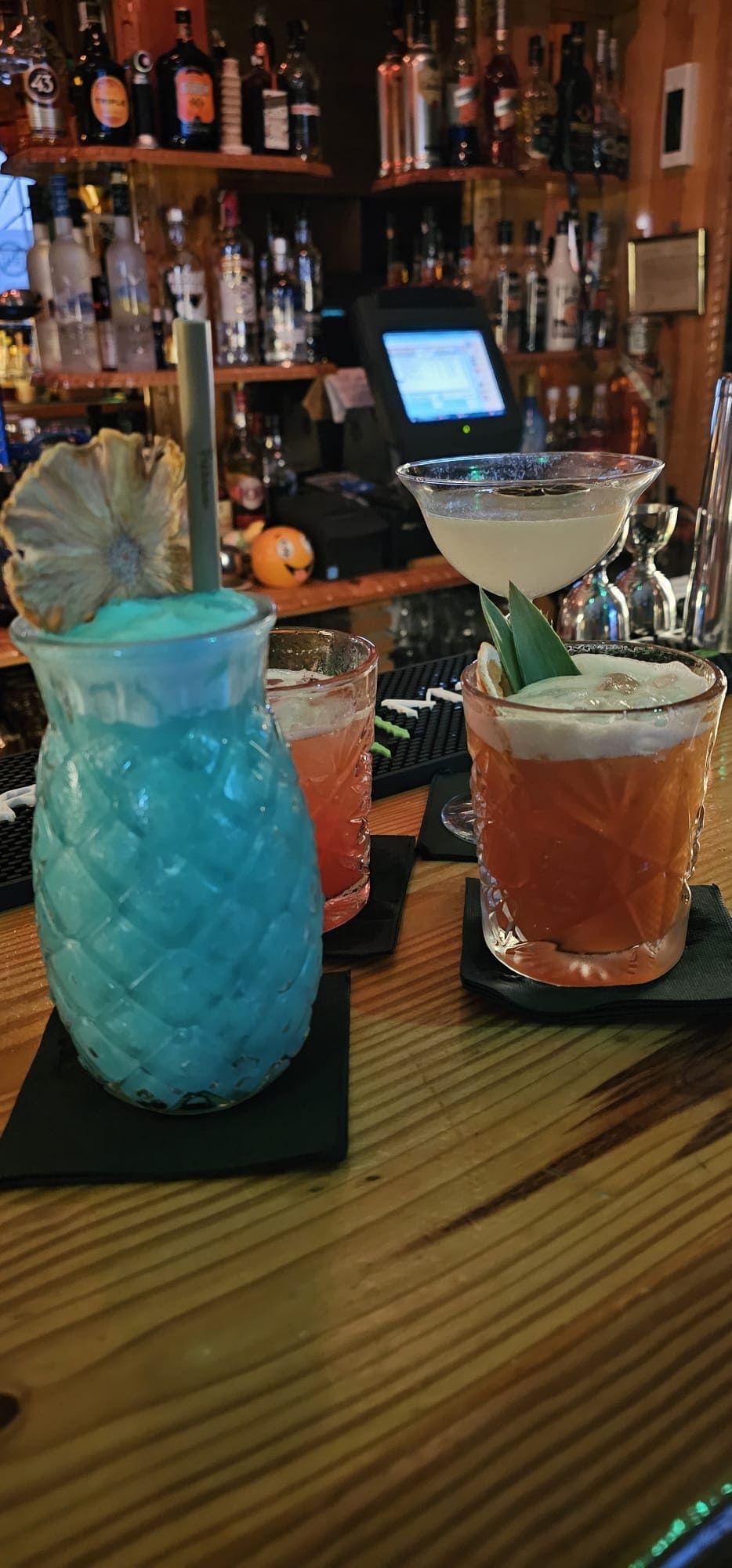 Tres cócteles sobre una barra de madera: uno azul, uno naranja con espuma y una bebida rosa más pequeña detrás de ellos.