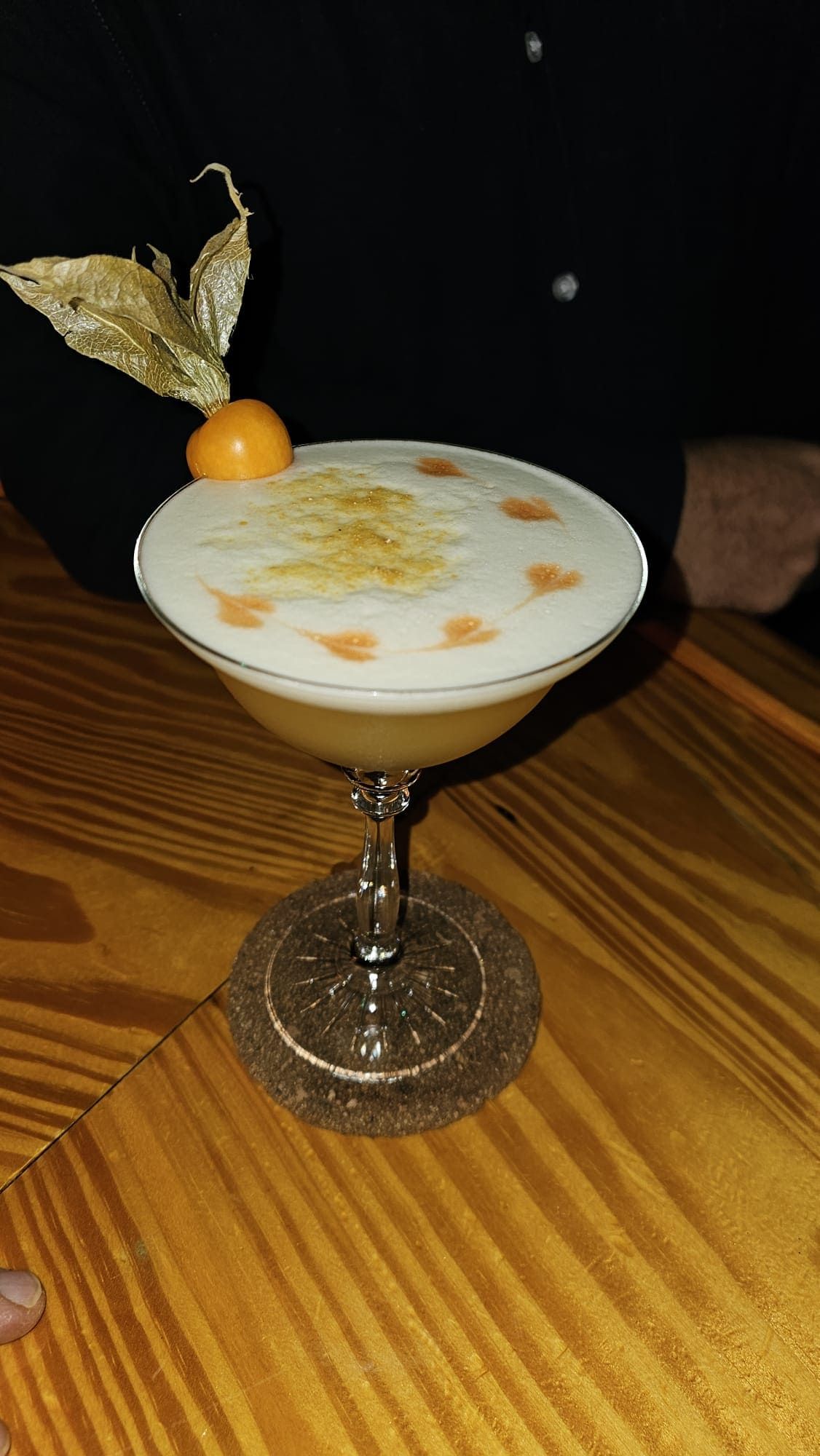 Un cóctel Pisco Sour en una copa con tallo, adornado con una baya de physalis y puntos de amargo, sobre una mesa de madera.