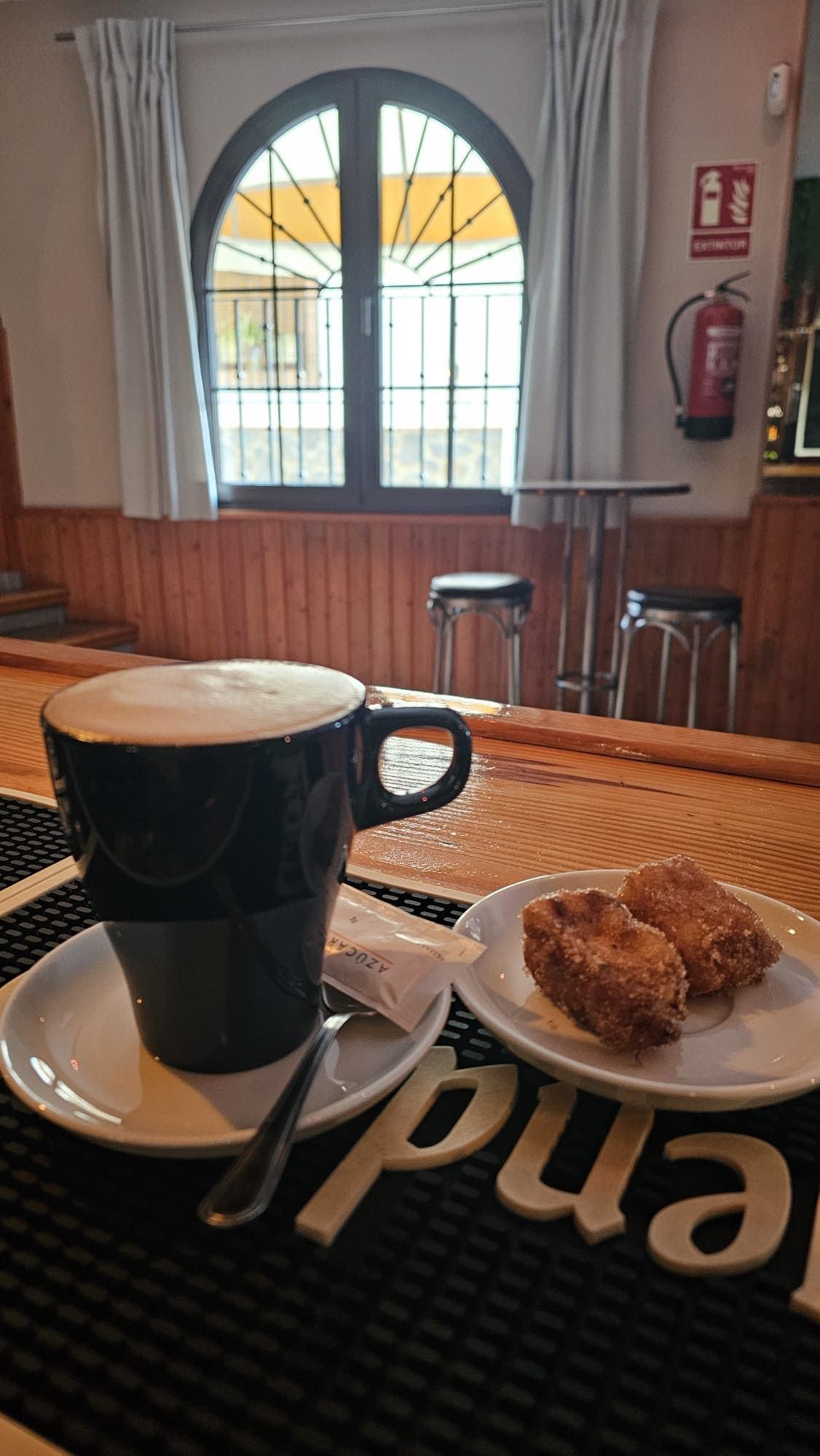 Una taza de café negra sobre un platillo, junto a un plato pequeño con dos pasteles en la barra de una cafetería.