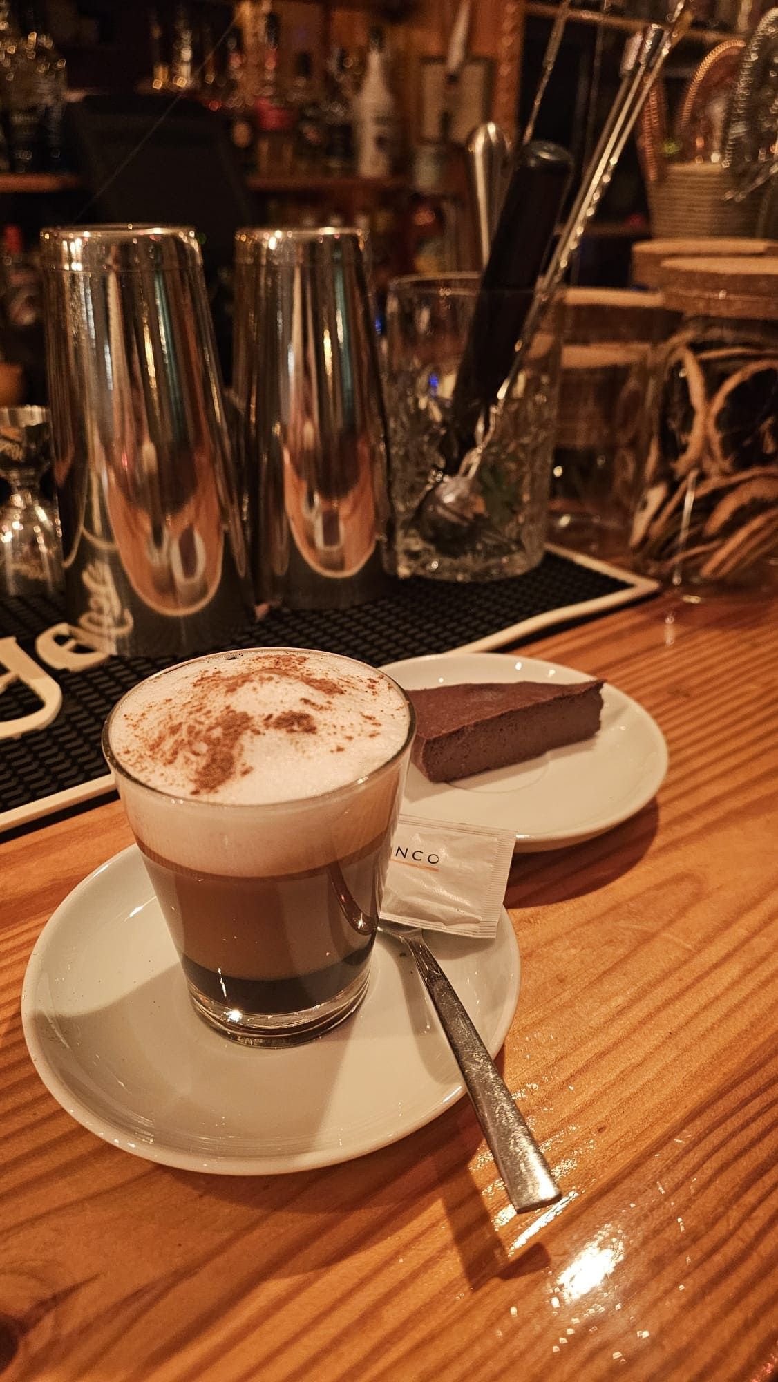 Una taza de café con leche espumada y canela, junto a una porción de pastel de chocolate sobre una barra de madera.