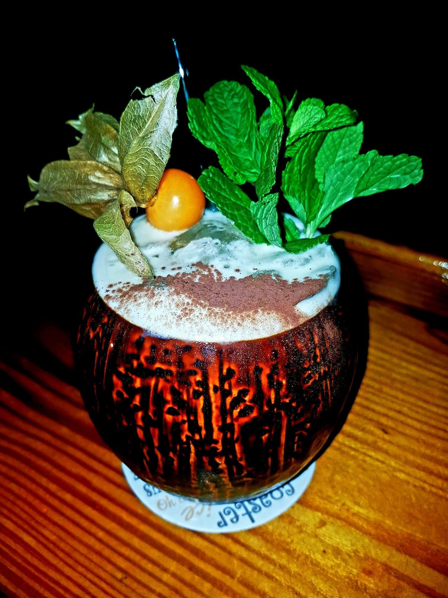 Un cóctel servido en una cáscara de coco oscura y texturizada, adornado con menta y una fruta de physalis, sobre una mesa de madera.