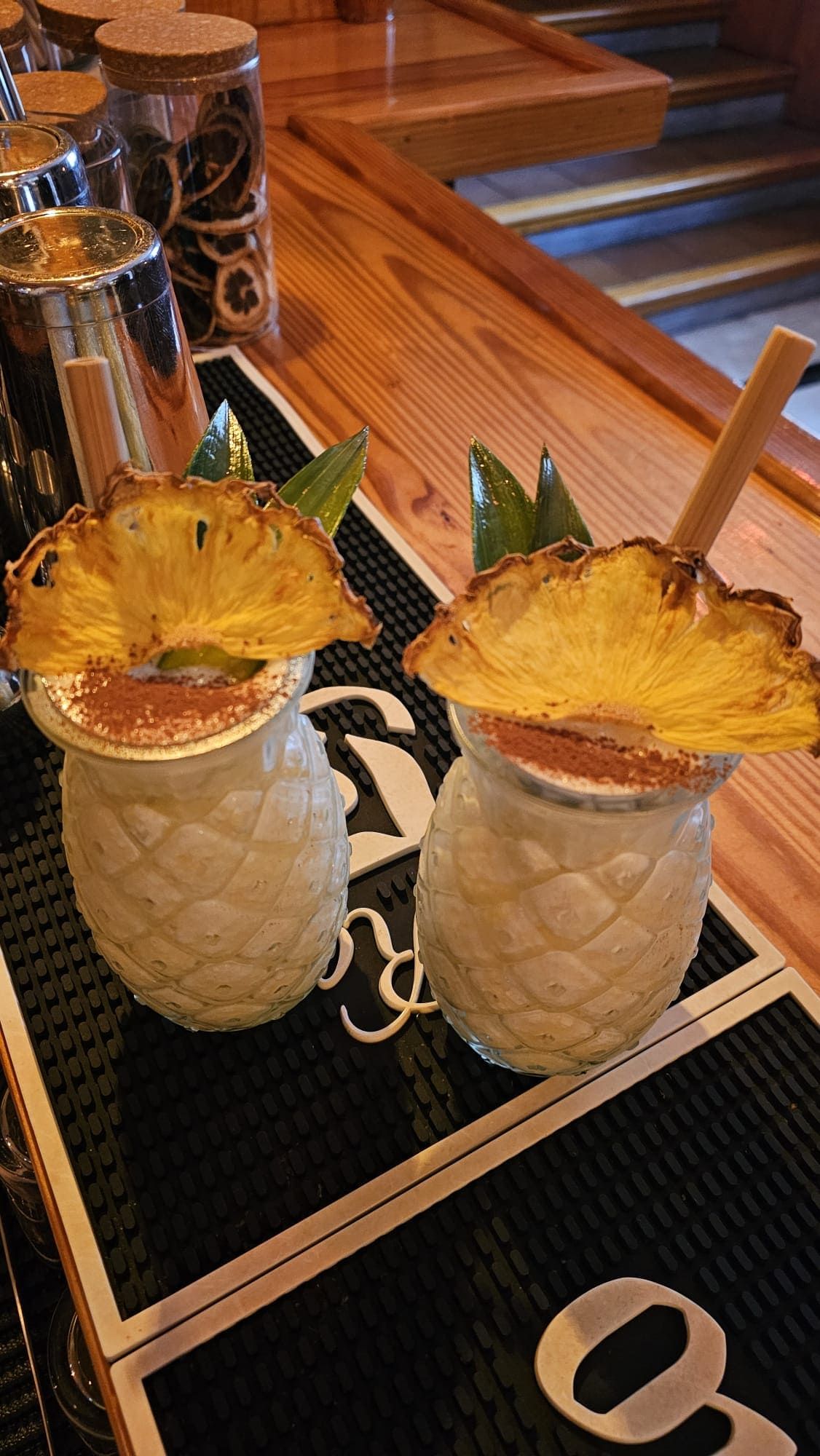 Dos piñas coladas en vasos con forma de piña, adornadas con pajitas y rodajas de piña, reposan sobre un mantel individual.