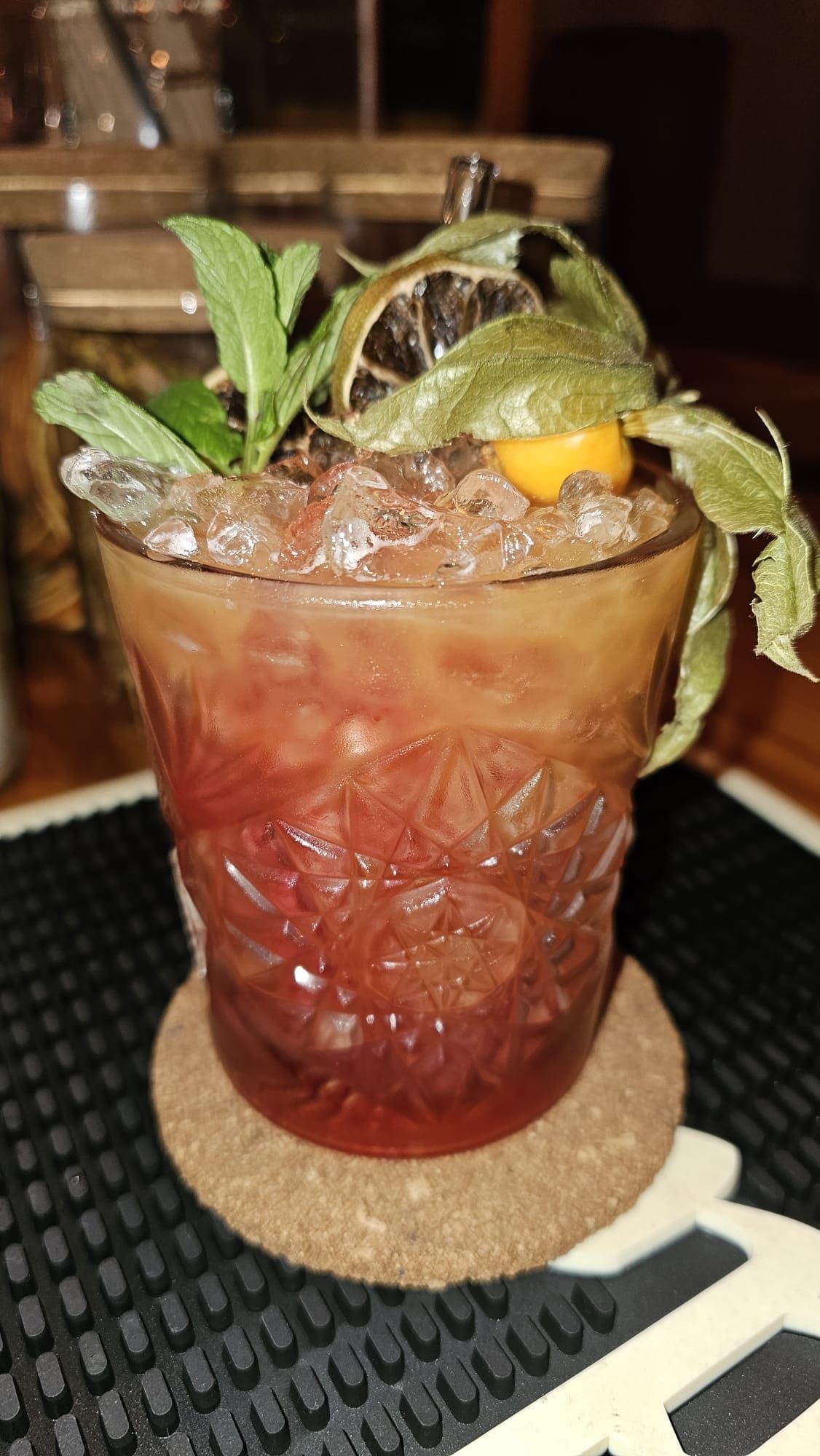 Un cóctel tropical y colorido con hielo picado, una ramita de menta, una rodaja de lima deshidratada y una uchuva sobre un posavasos de corcho.