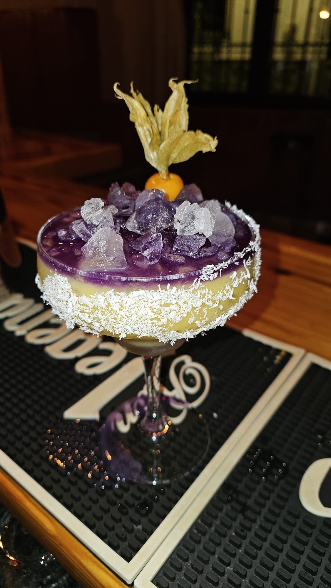Un cóctel servido en un vaso frío con el borde escarchado con coco rallado, coronado con un líquido morado, hielo picado y una baya dorada.
