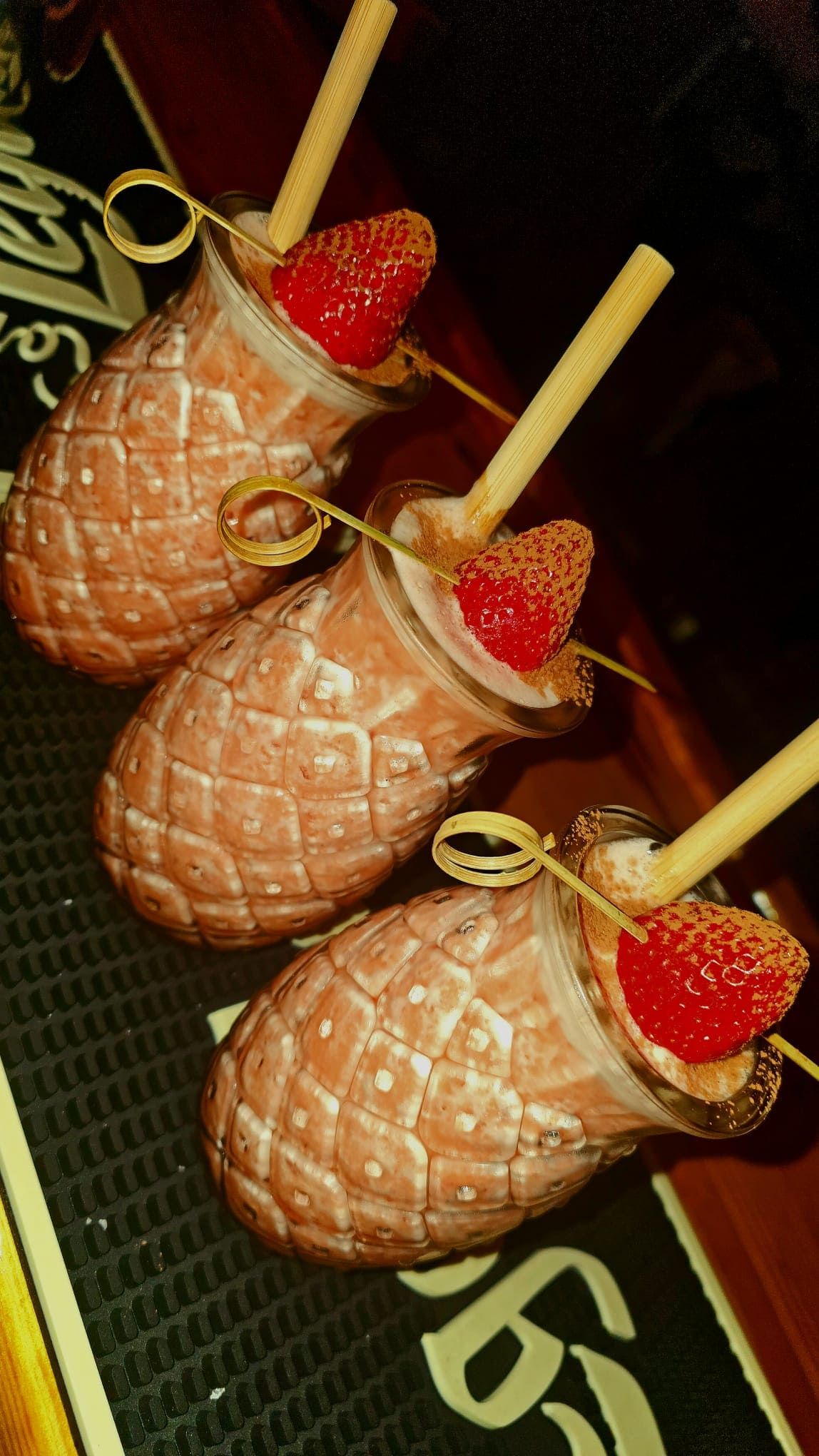 Tres vasos con forma de piña llenos de cócteles rosas, cada uno adornado con una fresa y una pajita sobre un mantel individual de bar.