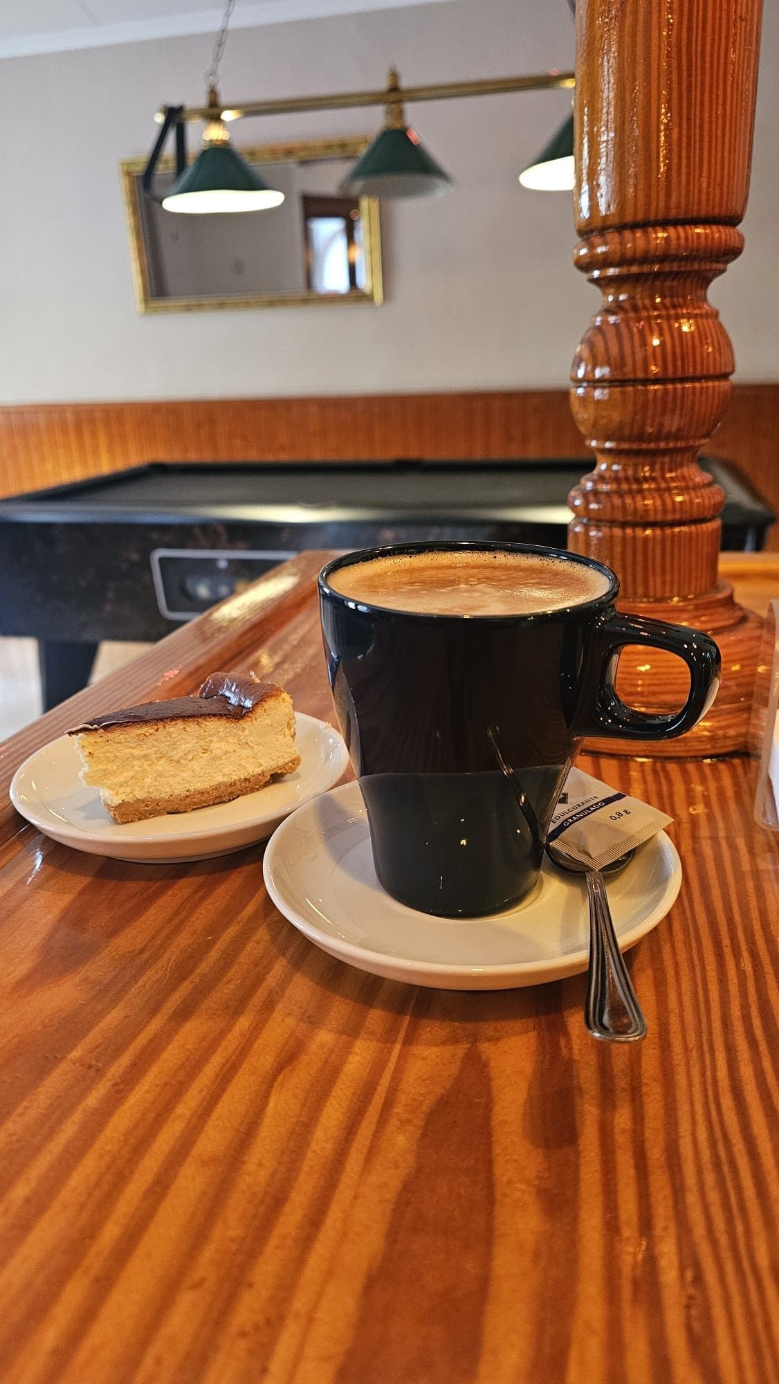 Un trozo de tarta de queso en un plato pequeño junto a una taza de café negra sobre una mesa de madera, con una mesa de billar al fondo.