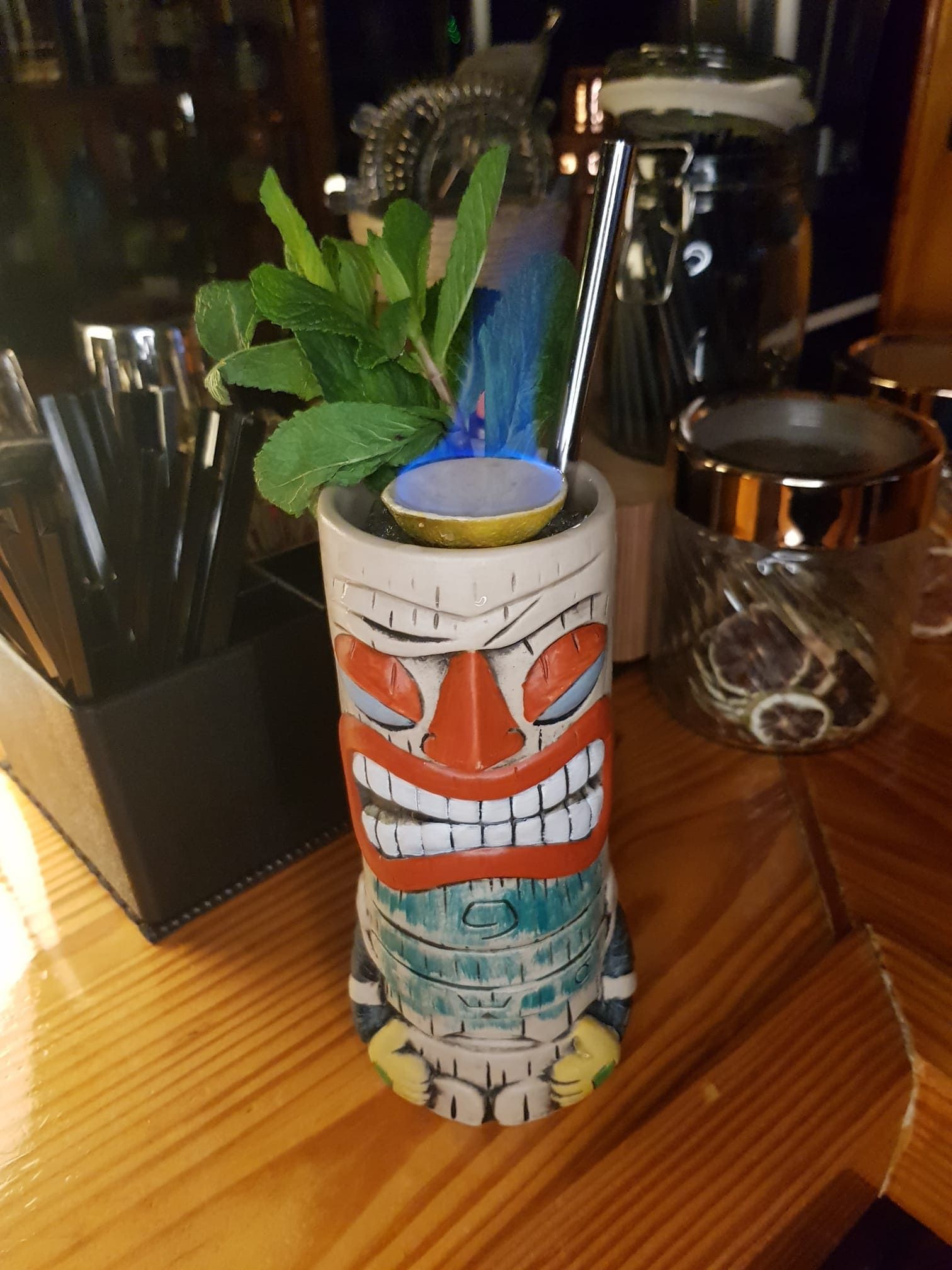Una taza Tiki en llamas, adornada con menta, reposa sobre una barra de madera junto a recipientes con ingredientes para cócteles.