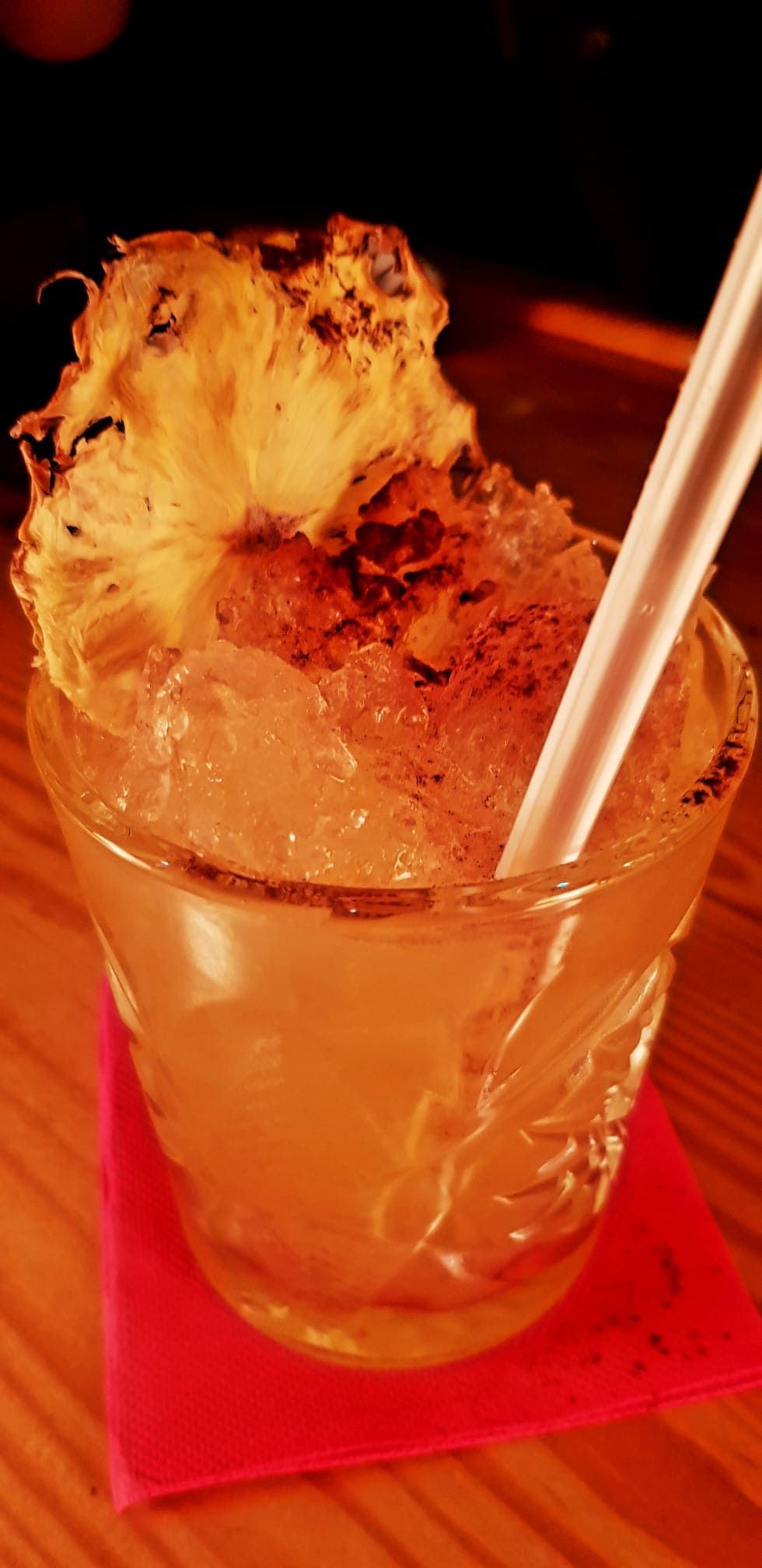 Un cóctel servido en un vaso estampado, adornado con una rodaja de piña deshidratada y lleno de hielo picado sobre una servilleta rosa.