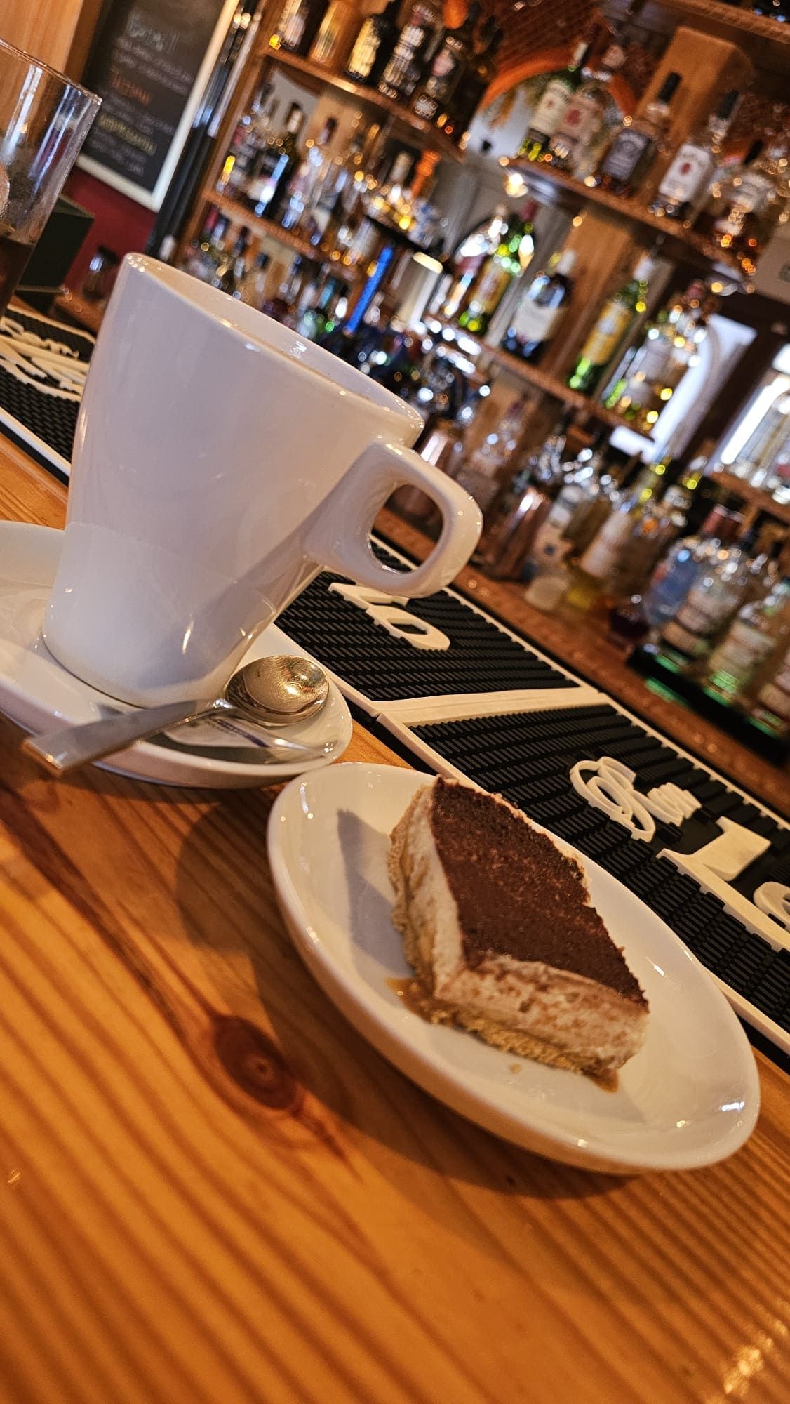 Una taza blanca y una porción de tiramisú en platos blancos, sobre una barra de madera frente a estantes llenos de botellas de licor.