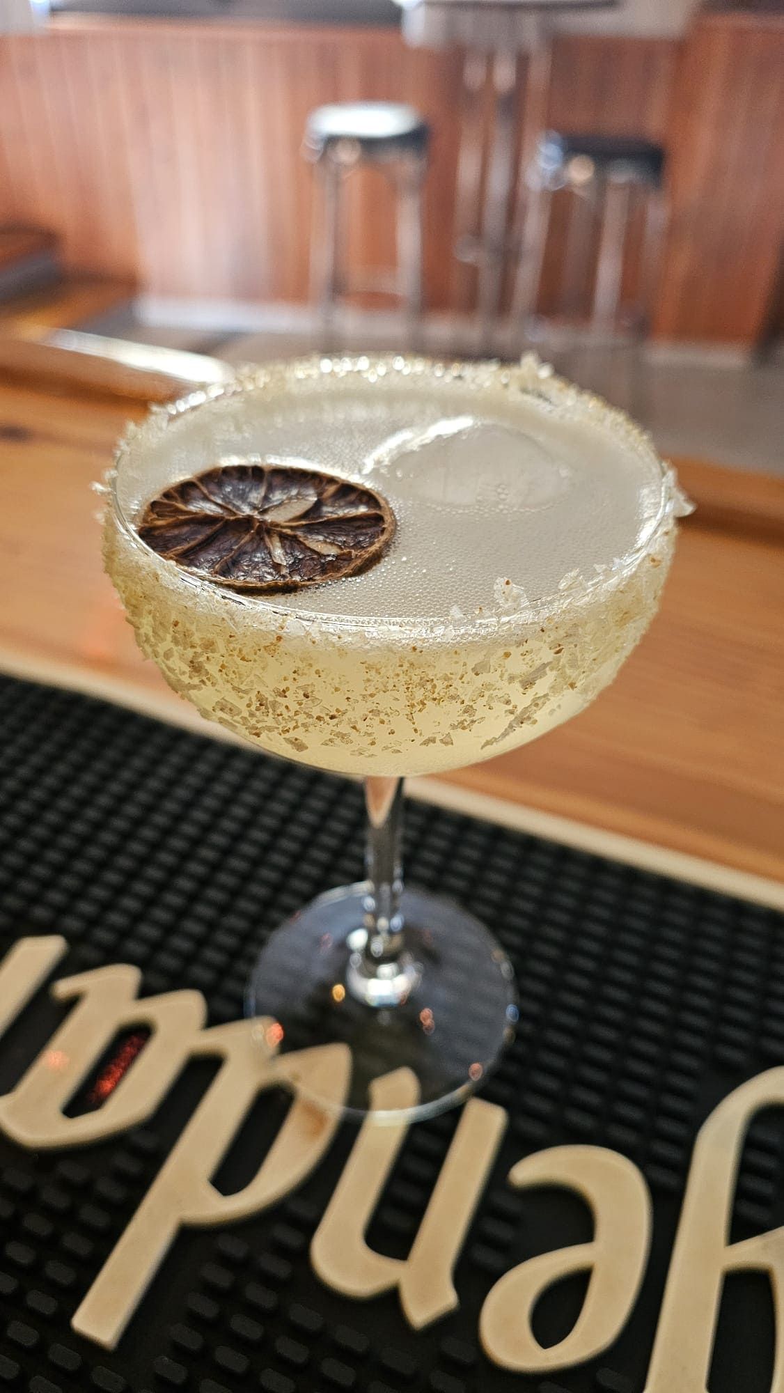 Un cóctel servido en una copa tipo coupe con el borde escarchado con azúcar, adornado con una rodaja de lima deshidratada, sobre un tapete de bar negro.